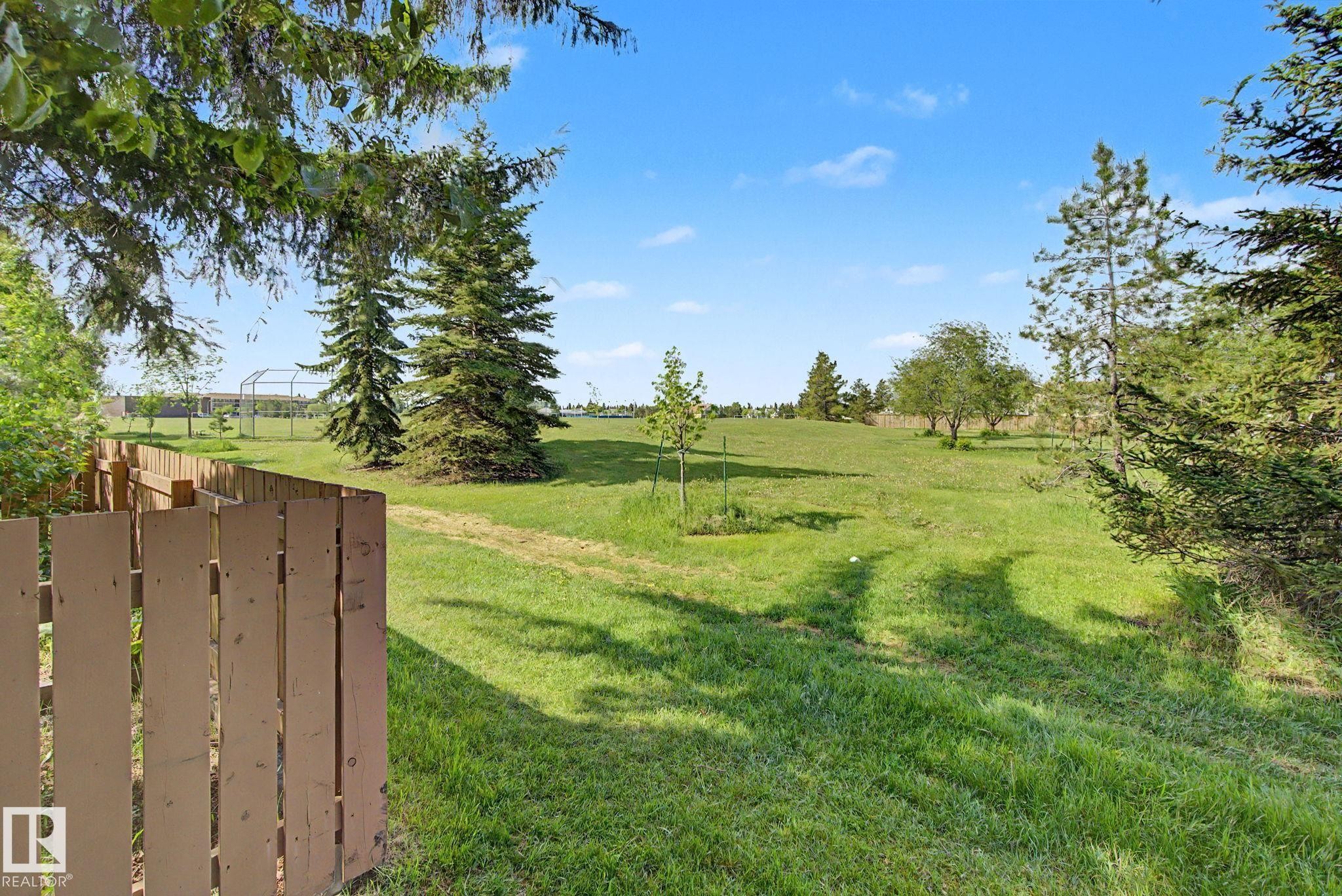 Expansive grassy area featuring a partial wood fence, mature evergreen trees, and scattered deciduous trees under a clear sky - #45 2020 105 St Nw, Edmonton, AB - Outdoor With View