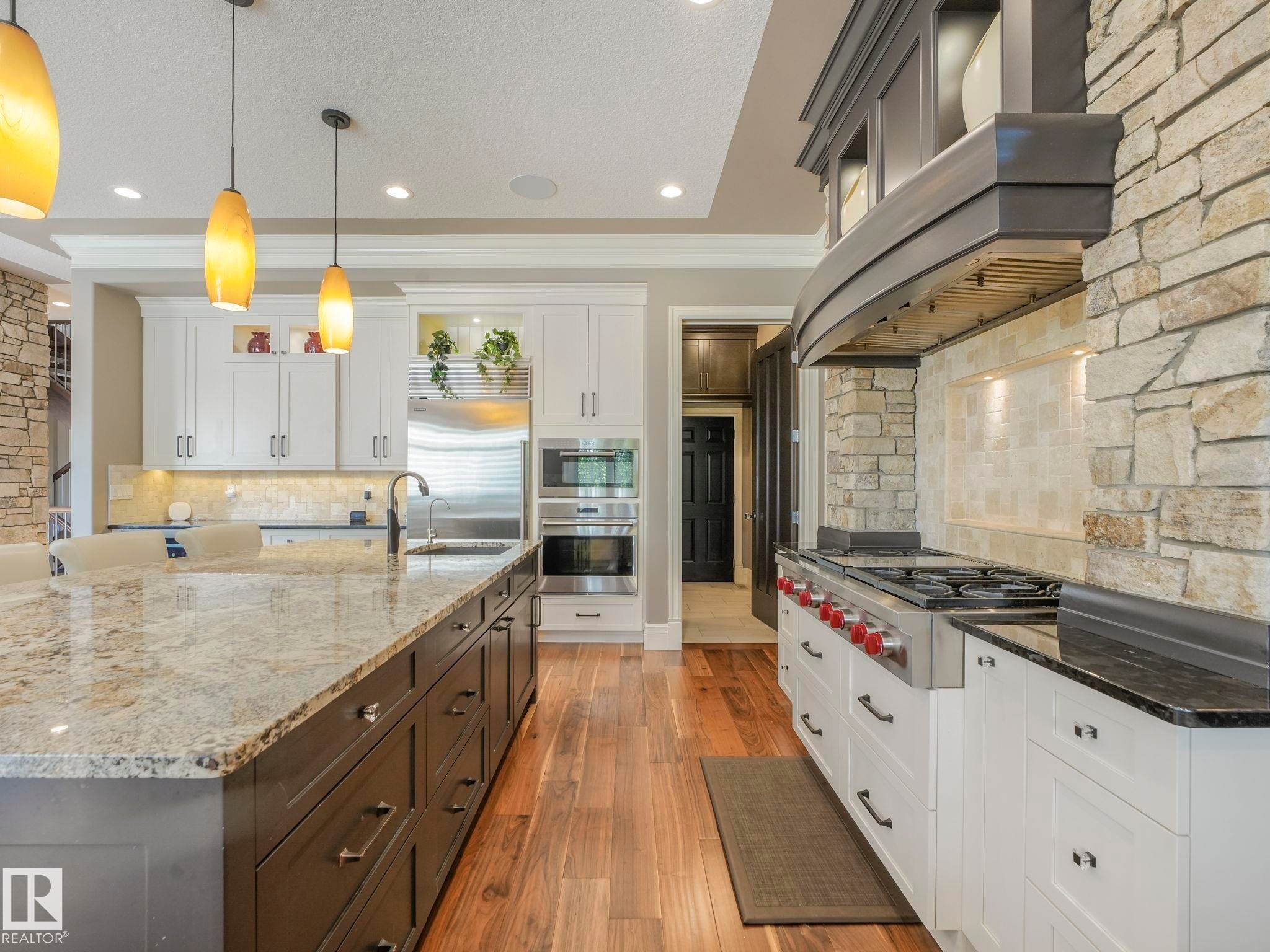 Gourmet kitchen featuring a stone accent wall, wood-finish flooring, a substantial kitchen island with a stone countertop, white cabinetry, and stainless steel appliances including a range and double oven - 2807 Wheaton Drive, Edmonton, AB