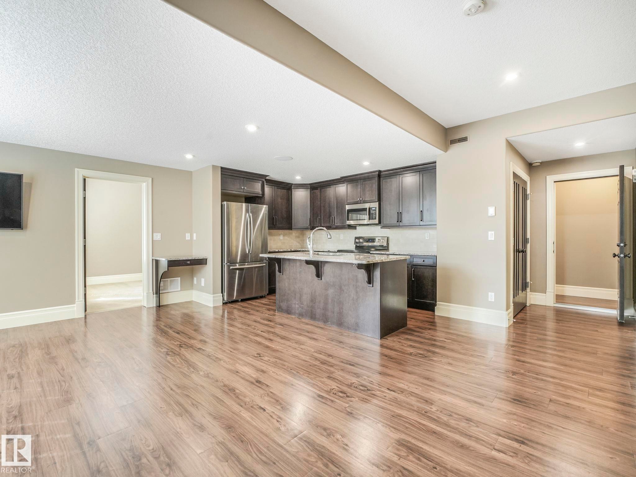 Open-concept kitchen and living area featuring wood-finish flooring, dark cabinetry, stainless steel appliances, a kitchen island with seating, and recessed lighting - 2807 Wheaton Drive, Edmonton, AB