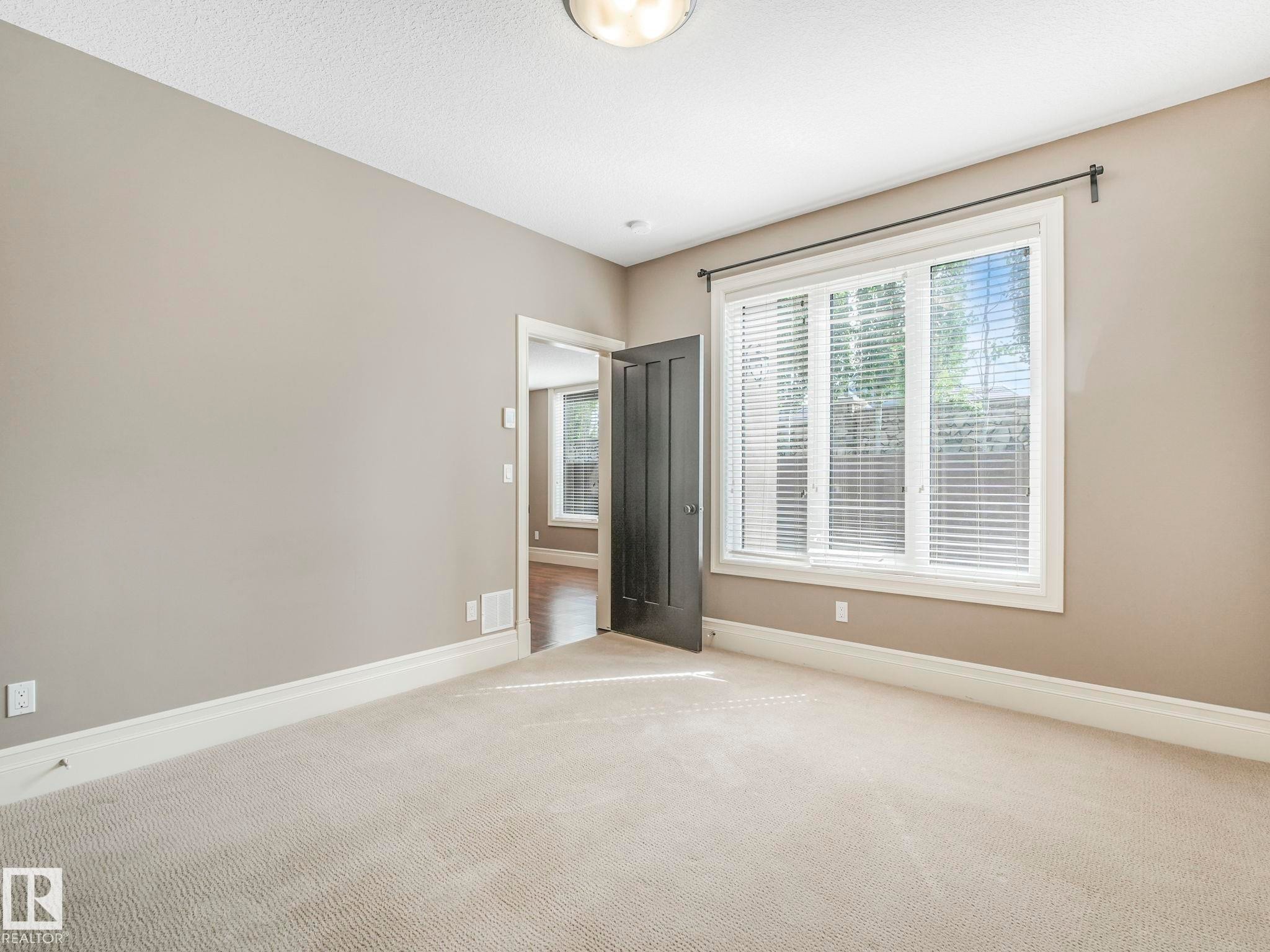 Carpeted room featuring a large window with white blinds, a dark paneled door, and neutral painted walls - 2807 Wheaton Drive, Edmonton, AB