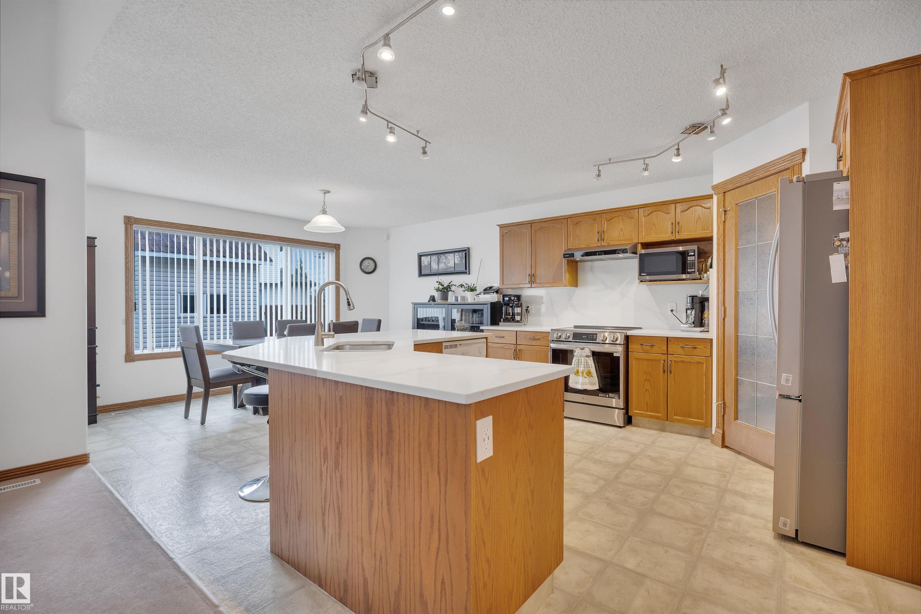 Open-concept kitchen featuring wood cabinetry, stainless steel appliances, a white countertop island with an integrated sink, and track lighting - 16512 87 Street Nw, Edmonton, AB - Indoor Photo Showing Kitchen
