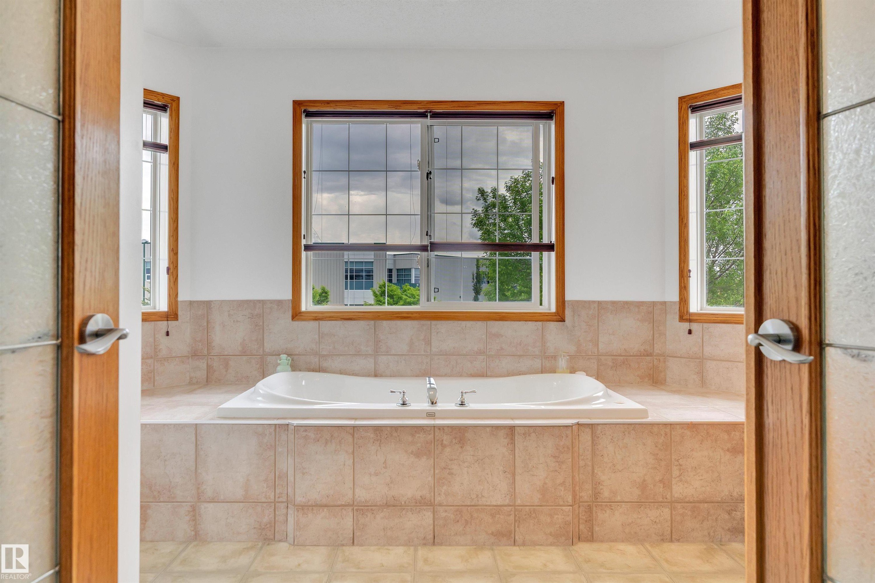 Built-in soaking tub with a tiled surround, three windows with wood-frame trim, and tile flooring - 16512 87 Street Nw, Edmonton, AB - Indoor Photo Showing Bathroom