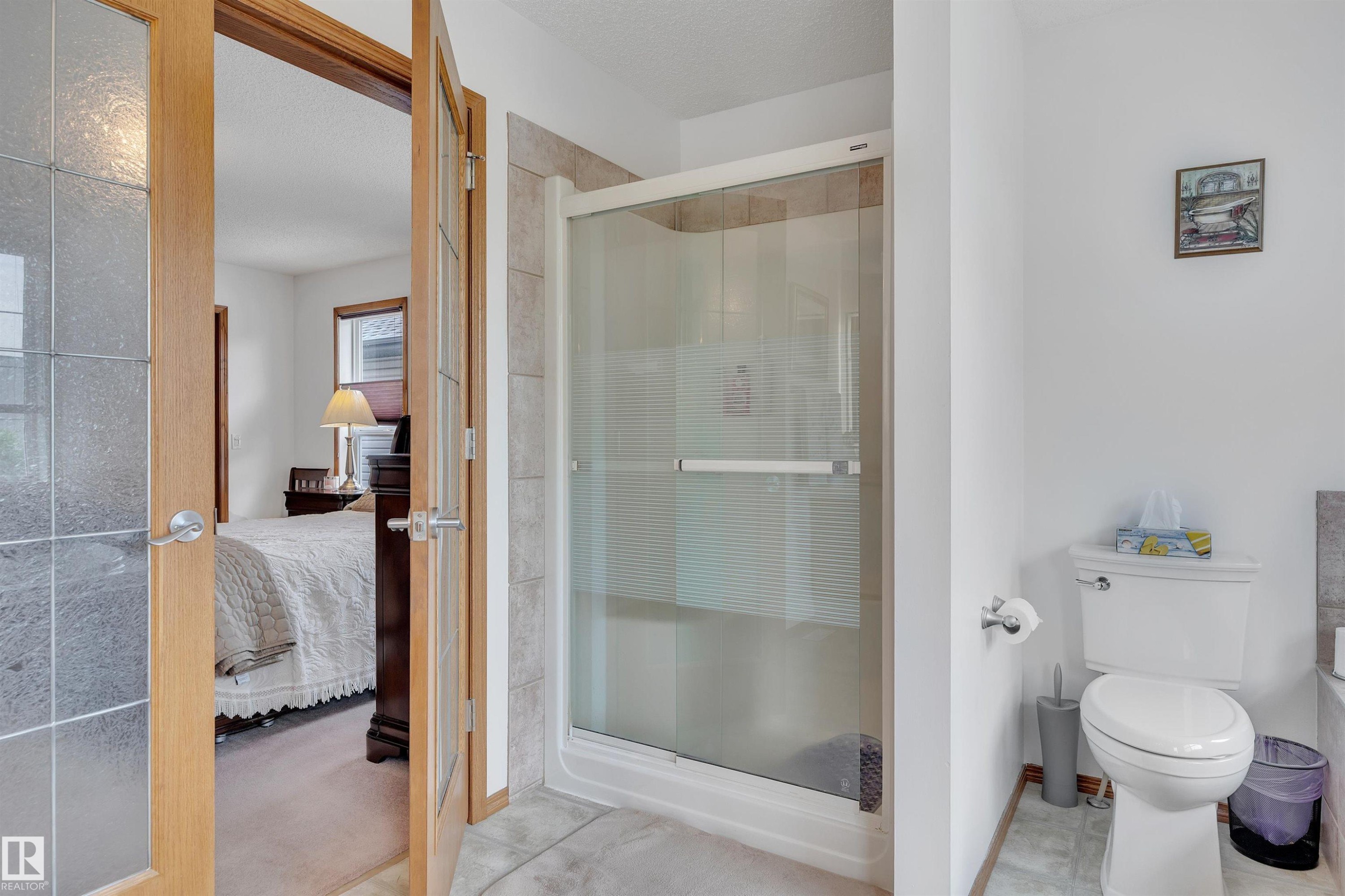 Glass-enclosed shower with a tiled surround, a white toilet, and light wood-finish French doors featuring textured glass panels - 16512 87 Street Nw, Edmonton, AB - Indoor Photo Showing Bathroom