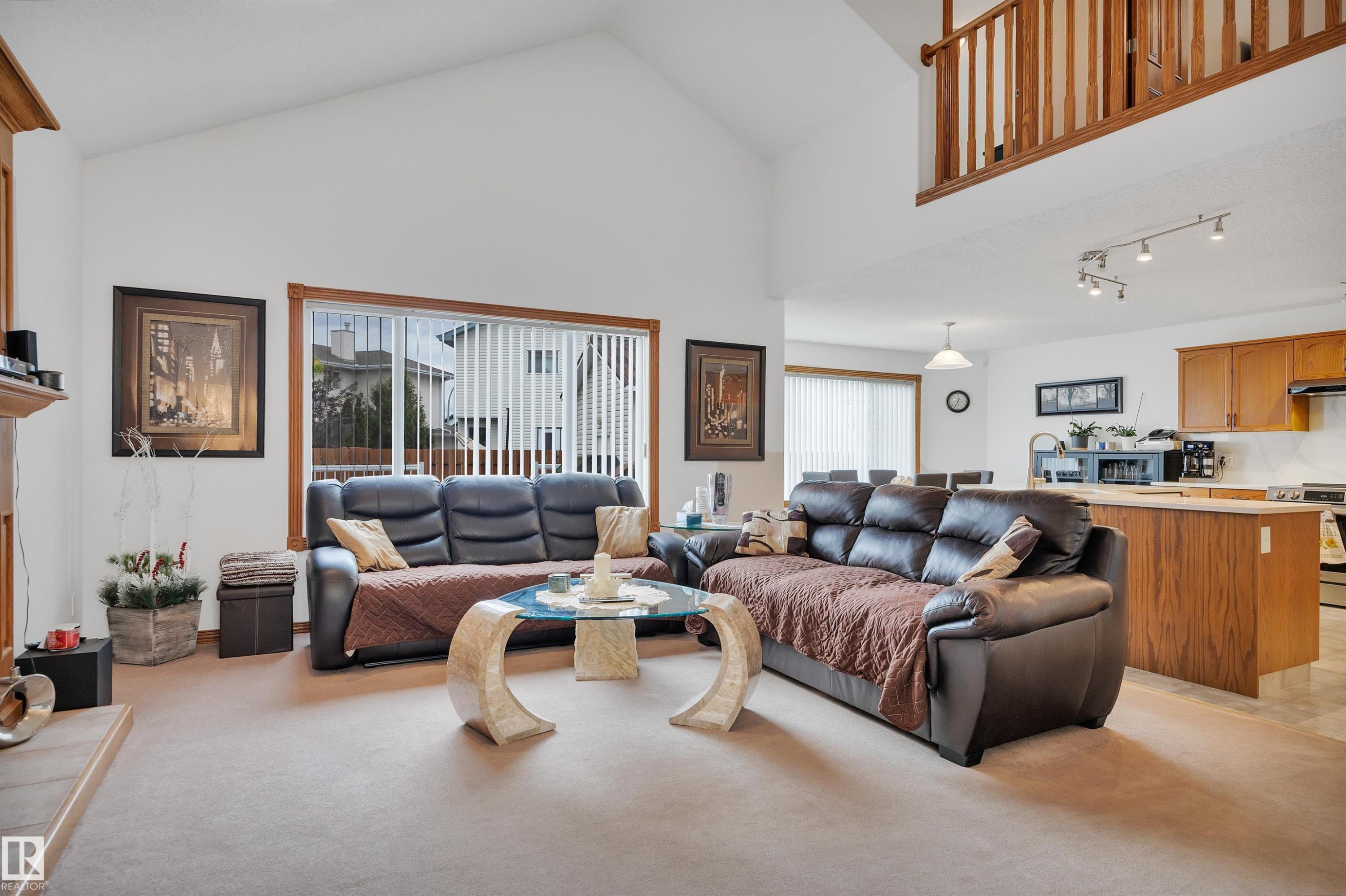 Open concept living space featuring vaulted ceilings, a large window with vertical blinds, a wood-finish railing, a kitchen with wood-finish cabinetry, and track lighting - 16512 87 Street Nw, Edmonton, AB - Indoor Photo Showing Living Room