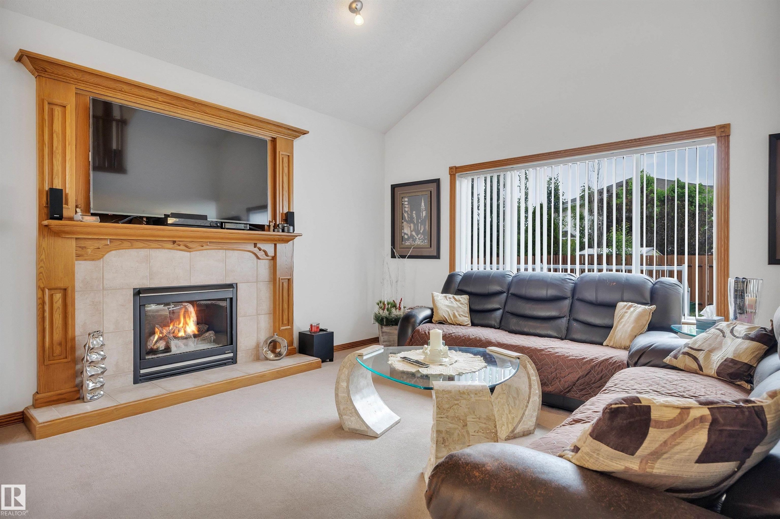 Living area with a vaulted ceiling, a wood-framed fireplace with a tile surround, and a large picture window with vertical blinds - 16512 87 Street Nw, Edmonton, AB - Indoor Photo Showing Living Room With Fireplace