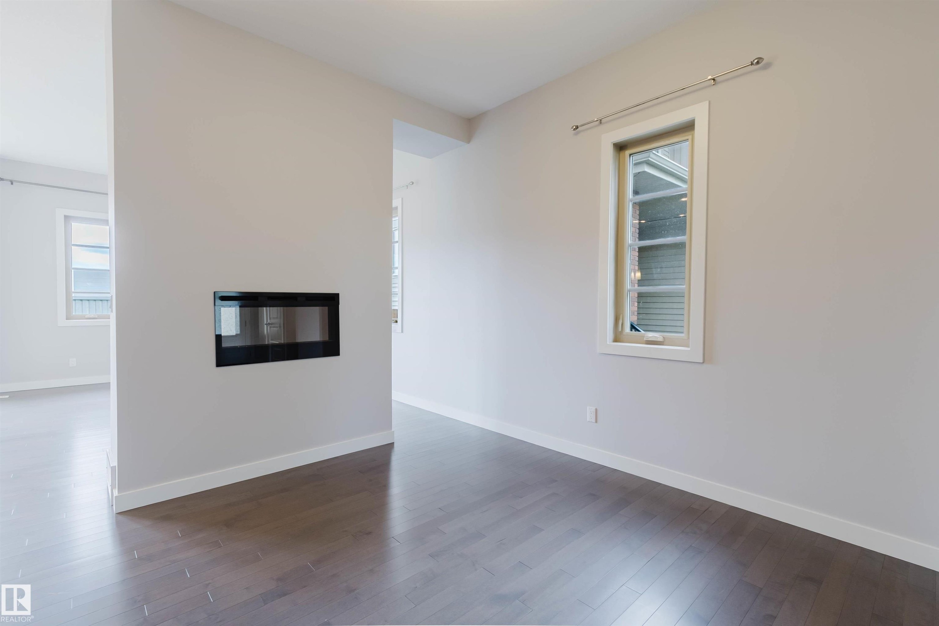 Modern linear fireplace integrated into a dividing wall, featuring dark wood-finish flooring, crisp white baseboards, light neutral wall paint, and a white-framed window - 2921 Anderson Crest Sw, Edmonton, AB - Indoor Photo Showing Other Room With Fireplace