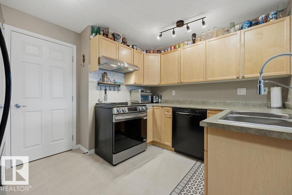 Kitchen featuring light wood cabinetry, stainless steel range with overhead ventilation, black dishwasher, and a double basin sink with a gooseneck faucet - 71 3040 Spence Wynd, Edmonton, AB