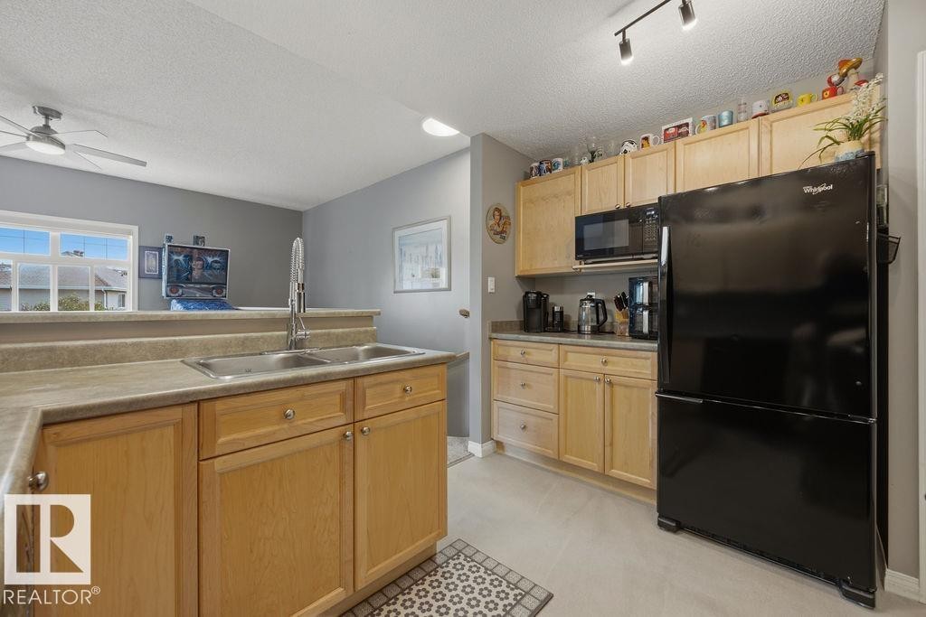 Kitchen featuring wood-finish cabinetry, a double basin stainless steel sink, black appliances, and track lighting - 71 3040 Spence Wynd, Edmonton, AB