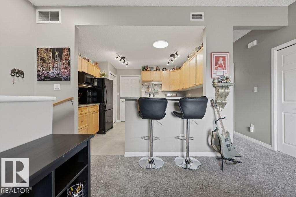 Open concept kitchen with a breakfast bar, light wood cabinetry, black appliances, and grey flooring - 71 3040 Spence Wynd, Edmonton, AB