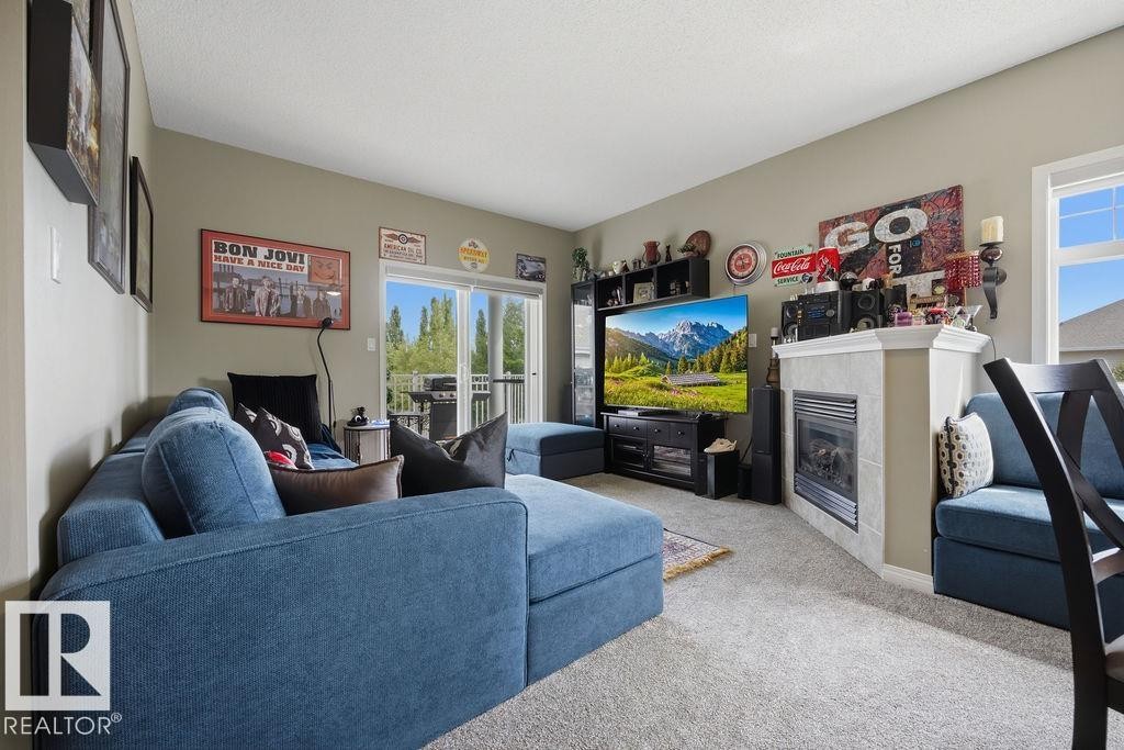 Living area featuring a tiled fireplace with mantel, sliding glass door access to an exterior deck, soft gray carpeting, and neutral wall tones - 71 3040 Spence Wynd, Edmonton, AB