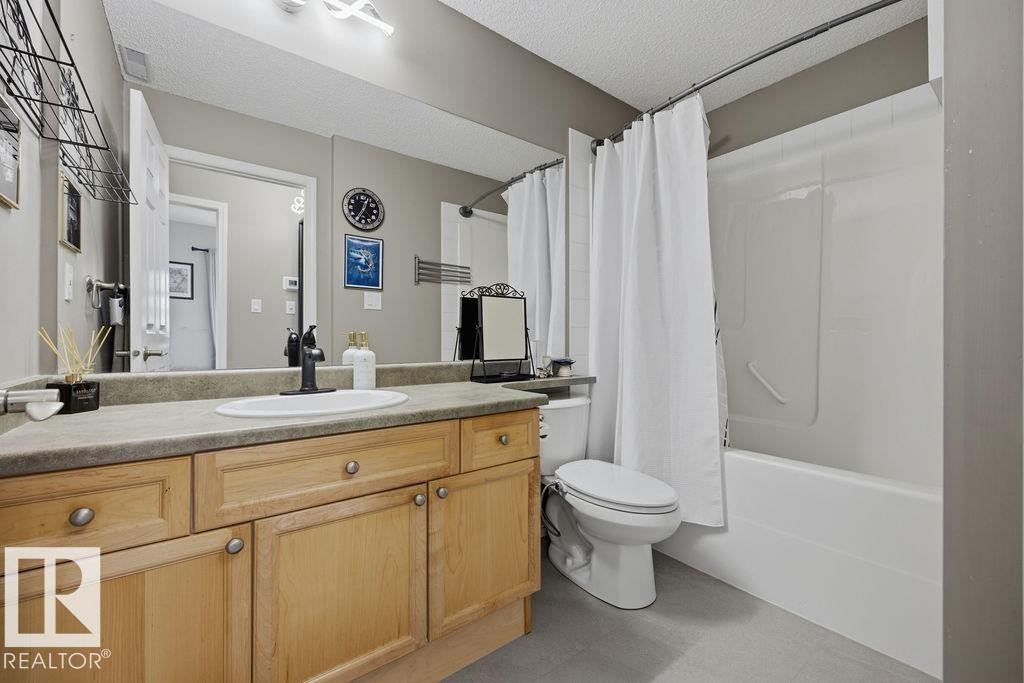 Bathroom featuring a light wood-finish vanity with a single basin sink, a large wall-mounted mirror, a toilet, and a combined bathtub and shower unit - 71 3040 Spence Wynd, Edmonton, AB