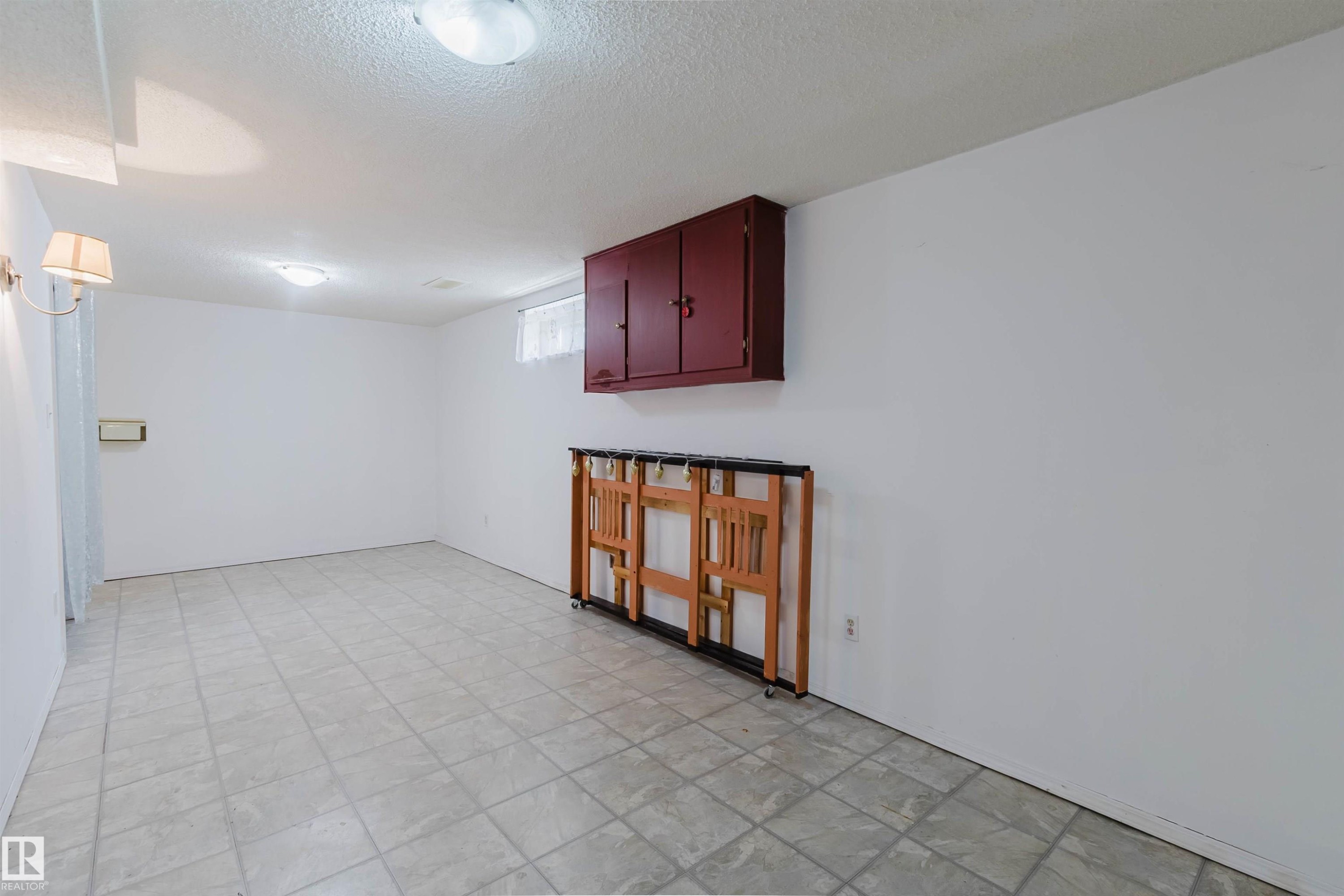 Spacious interior room featuring light-toned square tile flooring, a wall-mounted red wood-finish storage cabinet, a small window with light-colored curtains, and two ceiling-mounted dome lights providing illumination - 4 14310 80 Street, Edmonton, AB