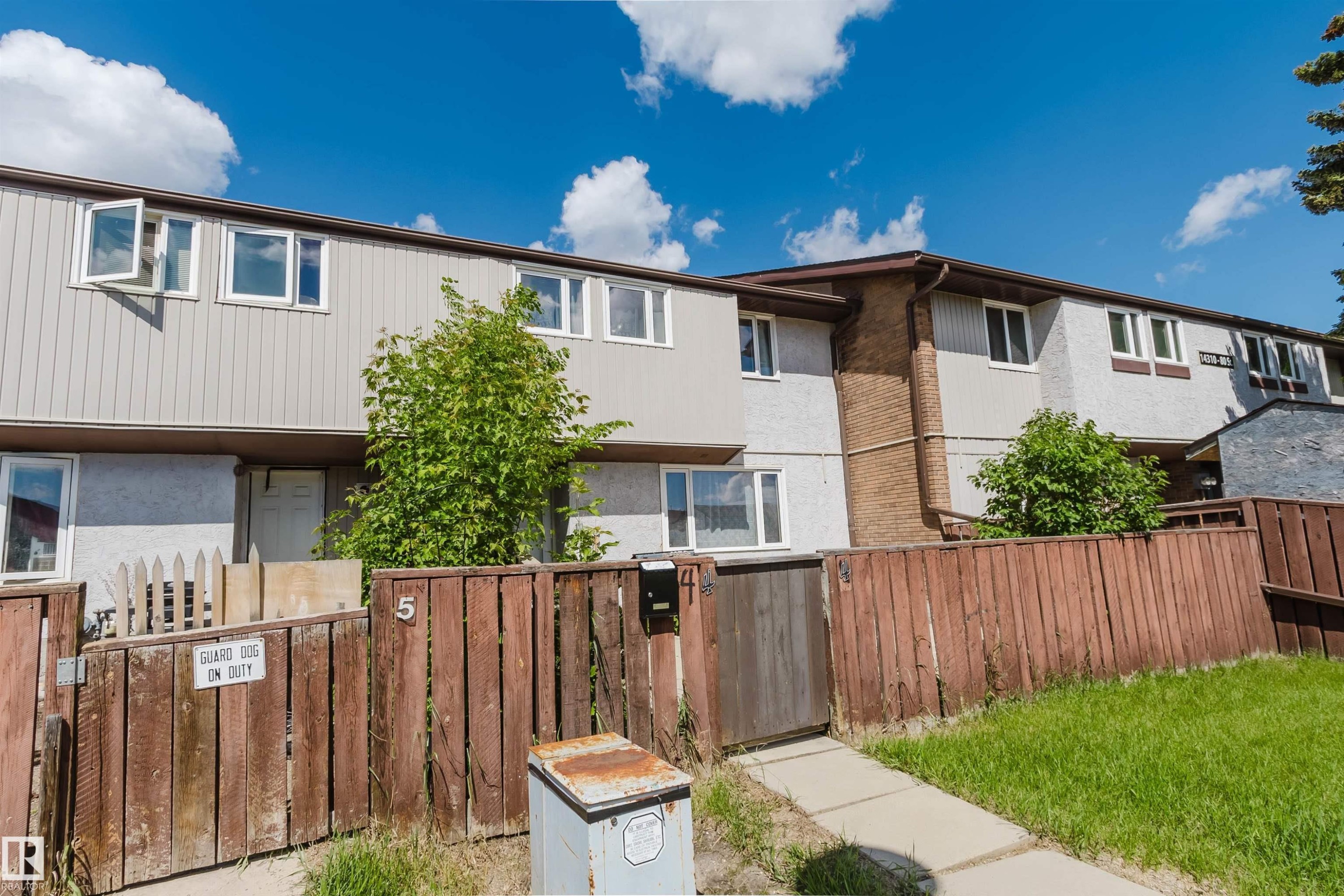 Two-story townhouse exterior featuring light-colored vertical siding and stucco finishes, multiple white-framed windows, and a brown wood fence with a pedestrian gate - 4 14310 80 Street, Edmonton, AB