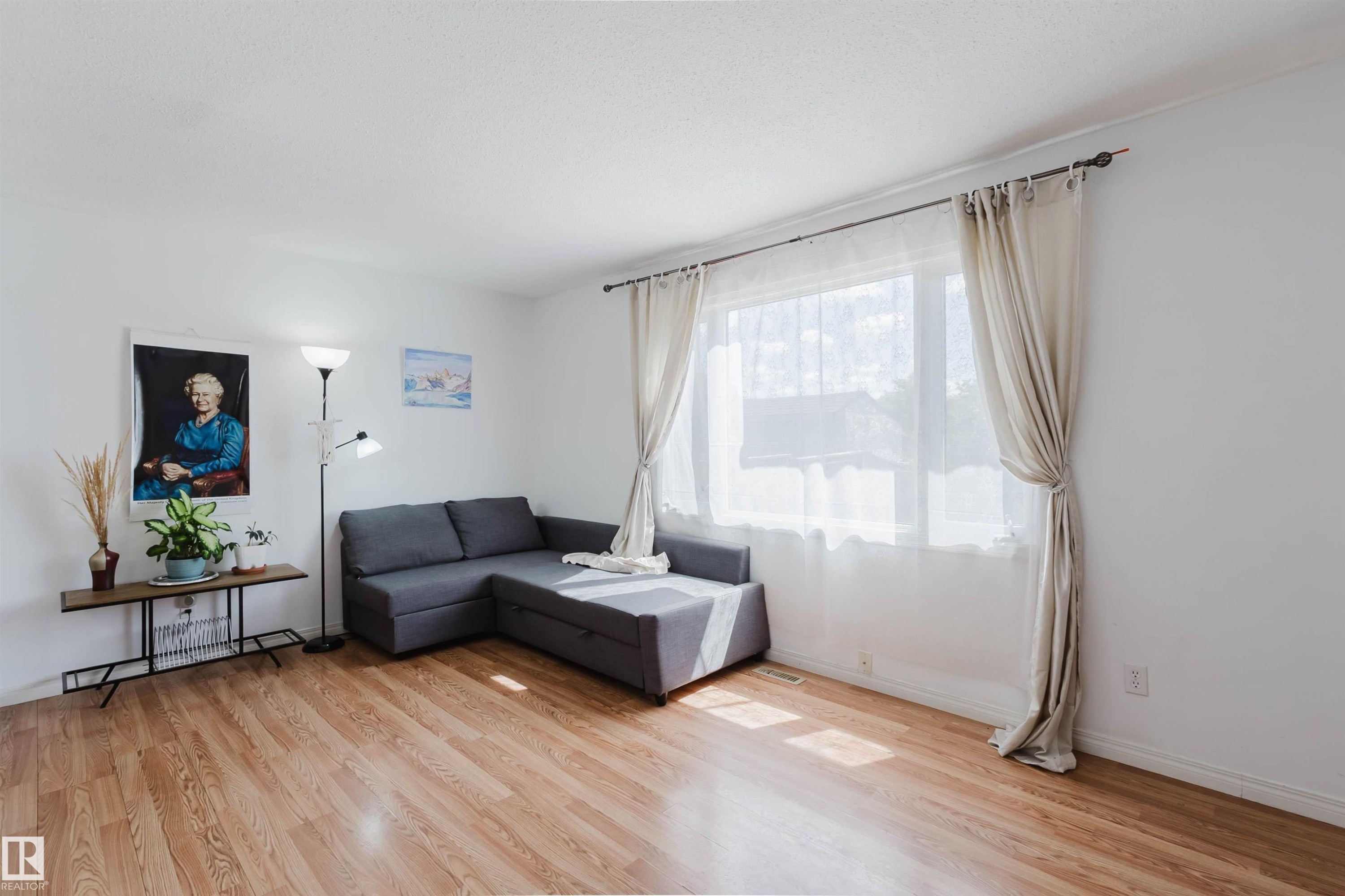 Spacious room featuring wood-finish flooring, white baseboards, and a large window with sheers and drapes - 4 14310 80 Street, Edmonton, AB