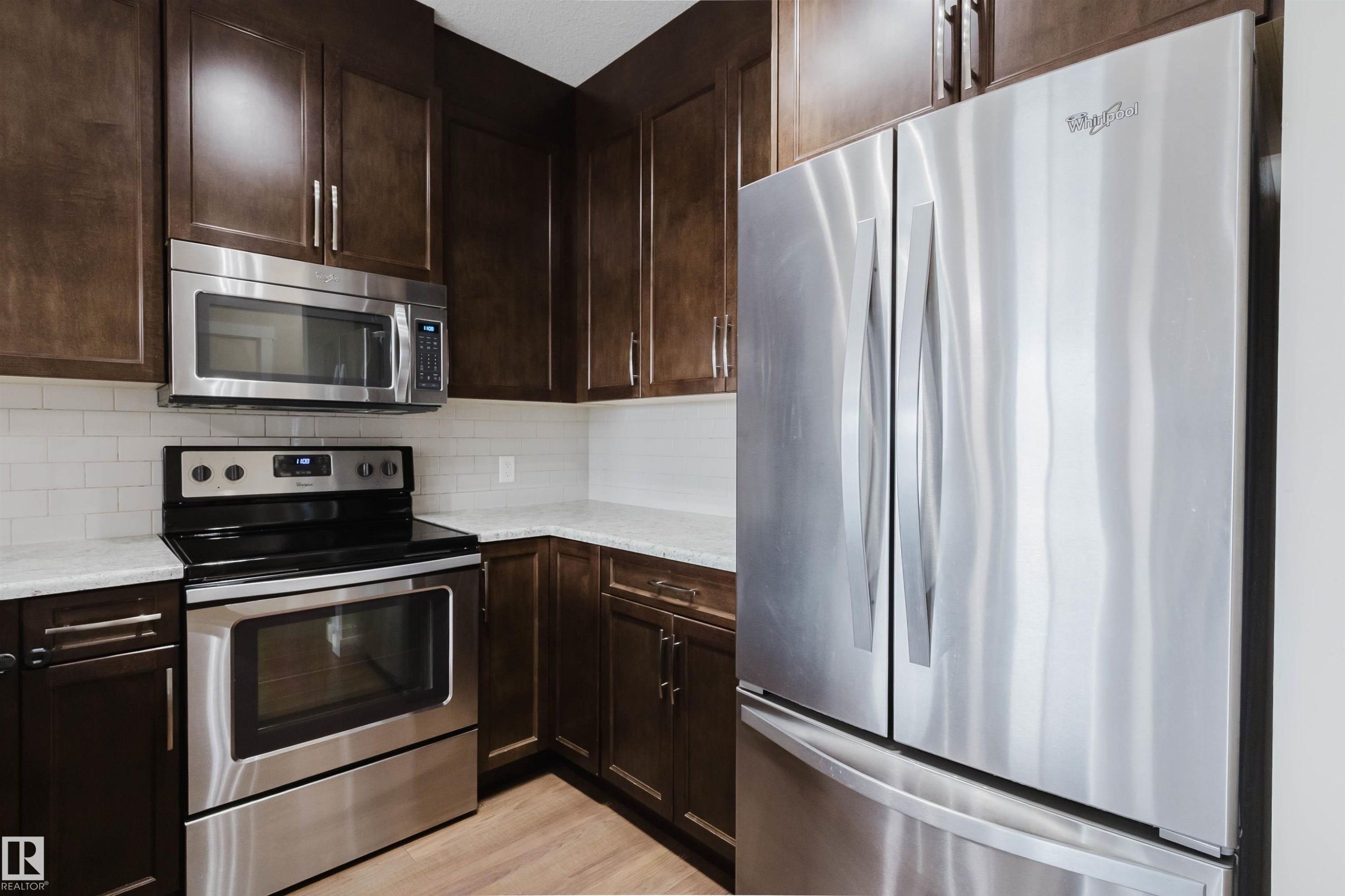 Kitchen featuring dark wood cabinetry, stainless steel appliances, light countertops, and a white subway tile backsplash - 402 3670 139 Avenue, Edmonton, AB