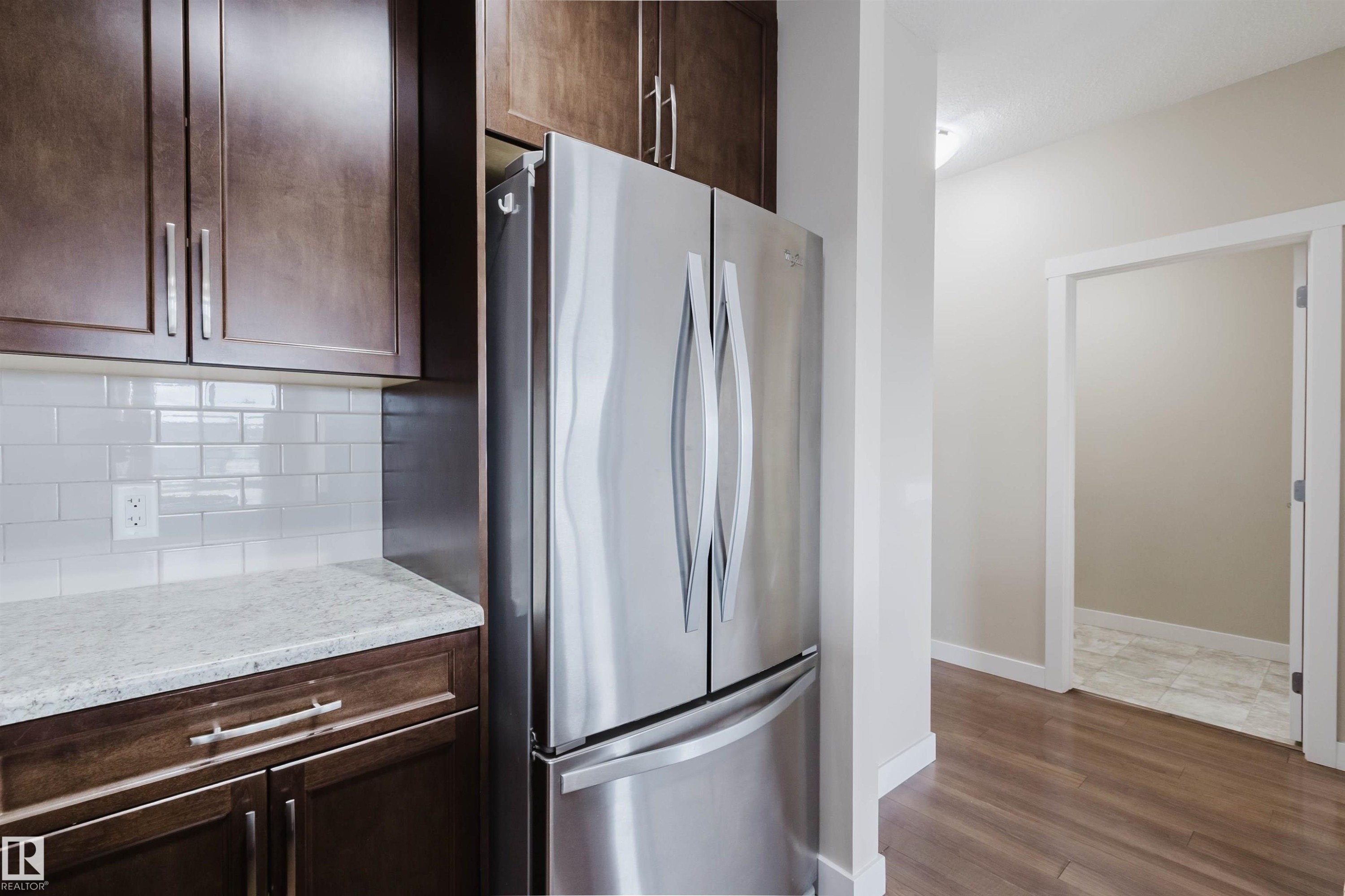 Stainless steel French-door refrigerator, rich wood-finish cabinetry with brushed nickel hardware, white subway tile backsplash, and light-toned solid surface countertops - 402 3670 139 Avenue, Edmonton, AB
