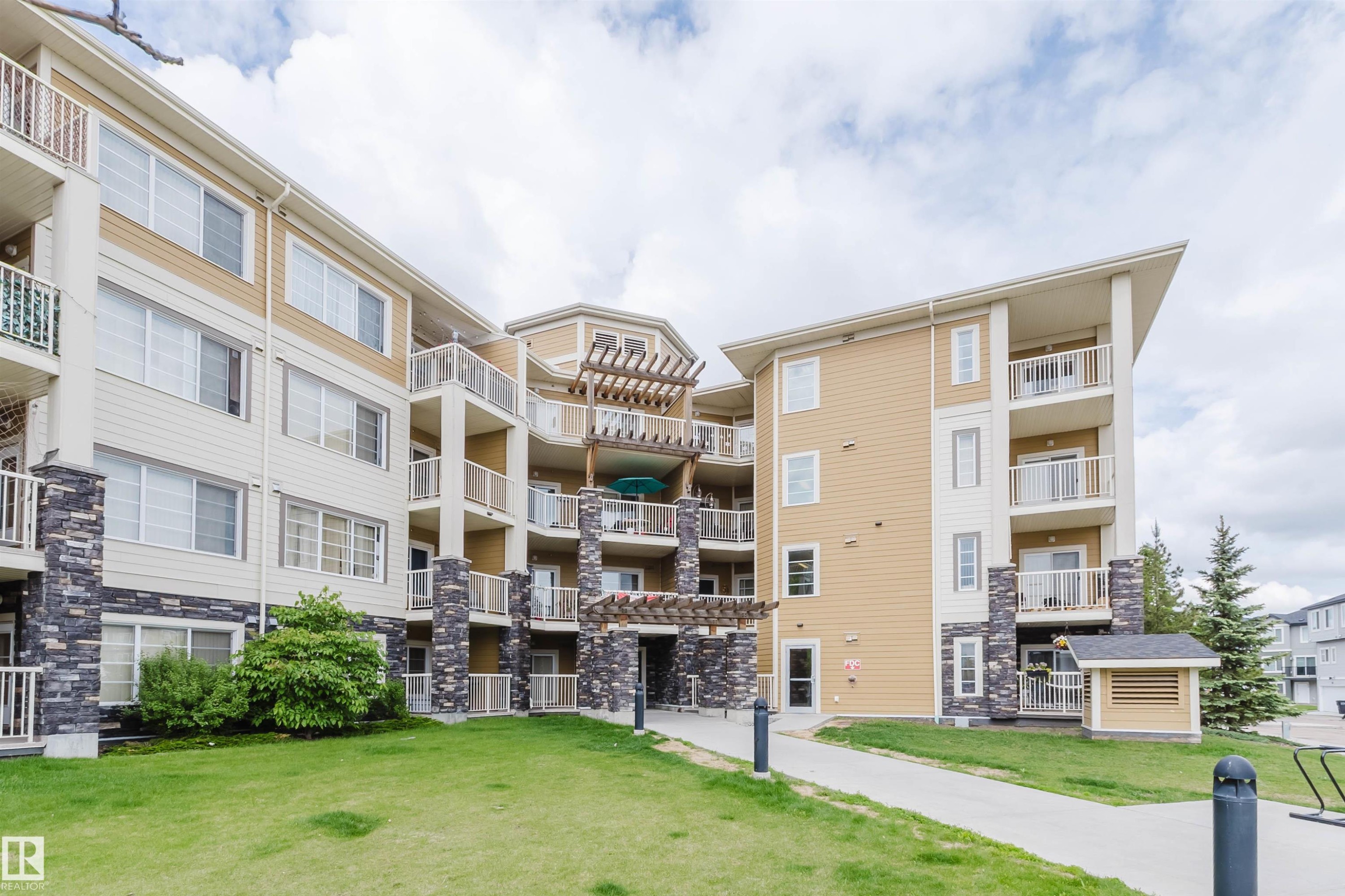 Multi-story building exterior featuring light-colored siding, stone accents, multiple balconies with metal railings, and a ground-level lawn area - 402 3670 139 Avenue, Edmonton, AB