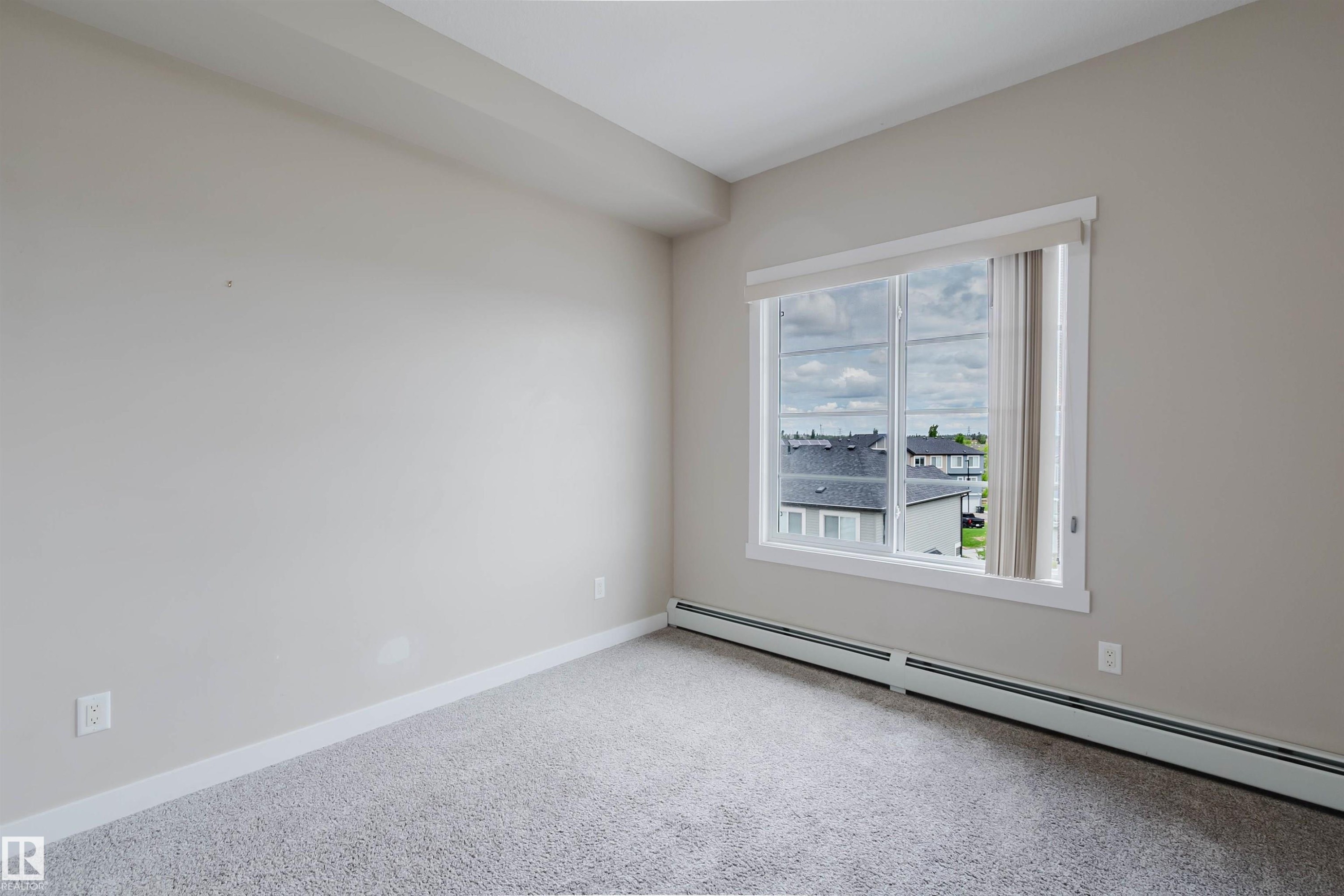 Carpeted interior space featuring a large window with white trim, light-colored walls, and baseboard heating - 402 3670 139 Avenue, Edmonton, AB