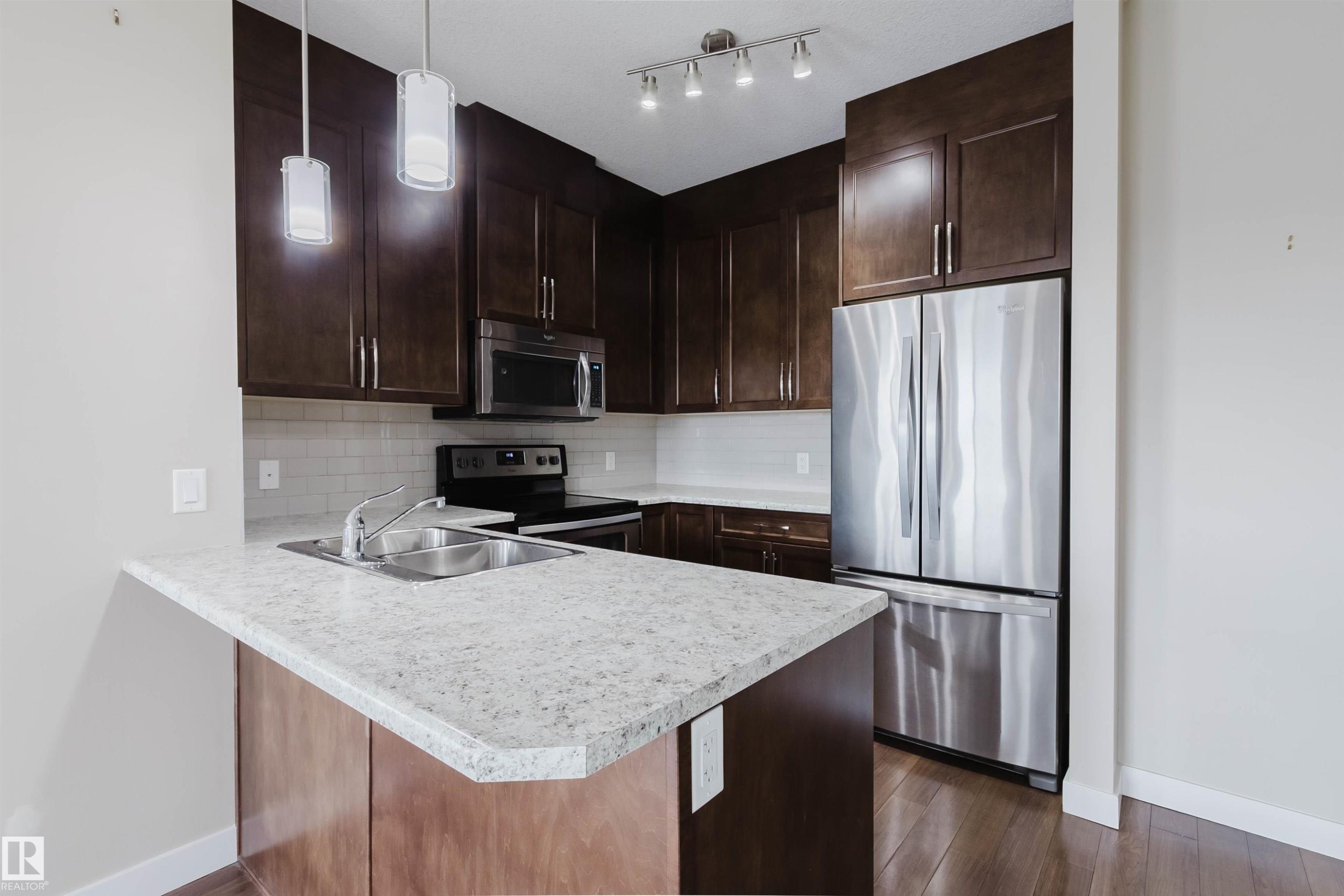 Kitchen featuring dark wood cabinetry, light speckled countertops, stainless steel appliances, and wood-finish flooring - 402 3670 139 Avenue, Edmonton, AB
