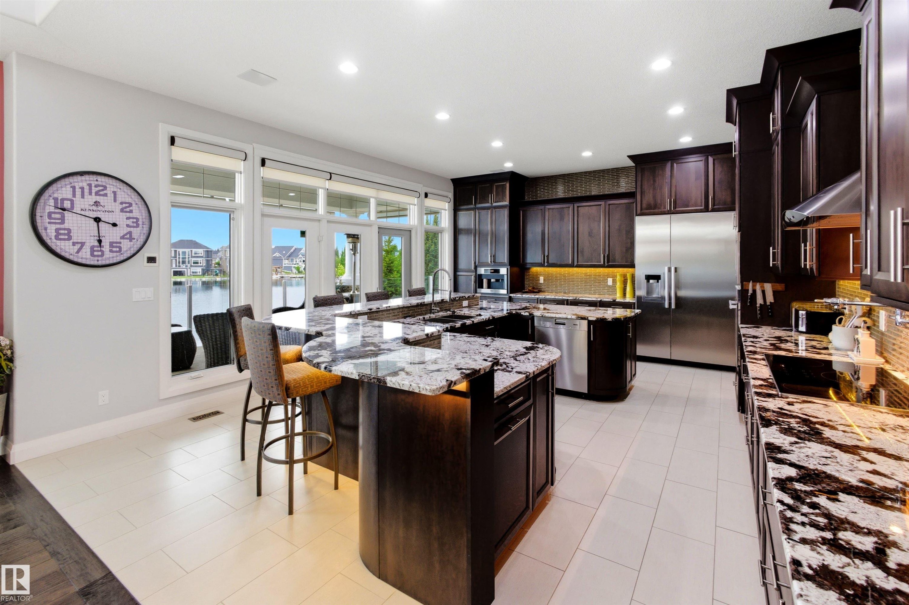 Expansive kitchen featuring dark wood cabinetry, a large kitchen island with a stone countertop, an integrated cooktop, stainless steel appliances, and white tile flooring - 9003 16 Avenue Sw, Edmonton, AB - Indoor Photo Showing Kitchen With Upgraded Kitchen