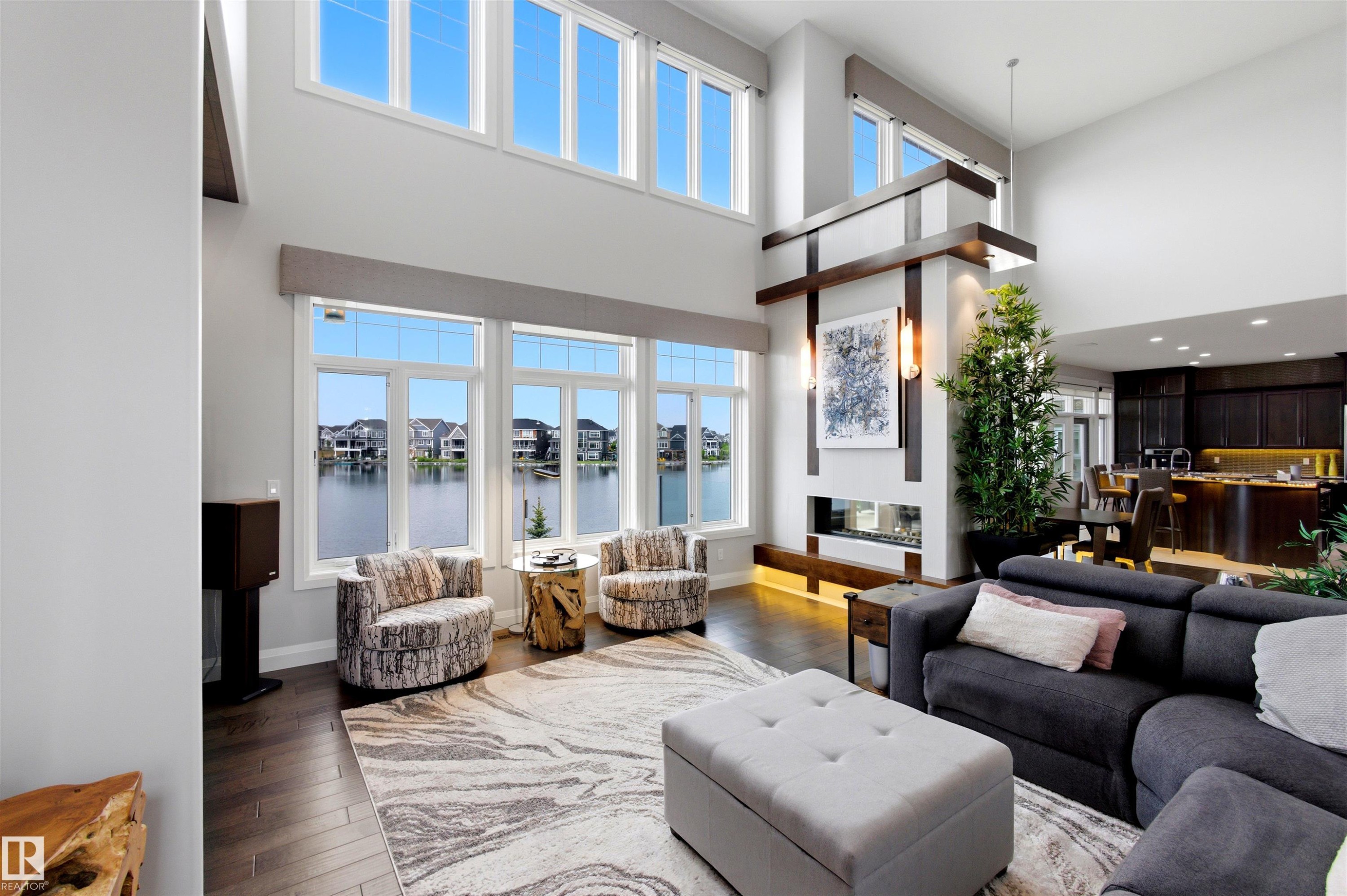 Two-story living area featuring large windows with waterfront exposure, dark wood-finish flooring, a modern linear fireplace with architectural wood accents, and a contemporary kitchen with dark cabinetry - 9003 16 Avenue Sw, Edmonton, AB - Indoor Photo Showing Living Room