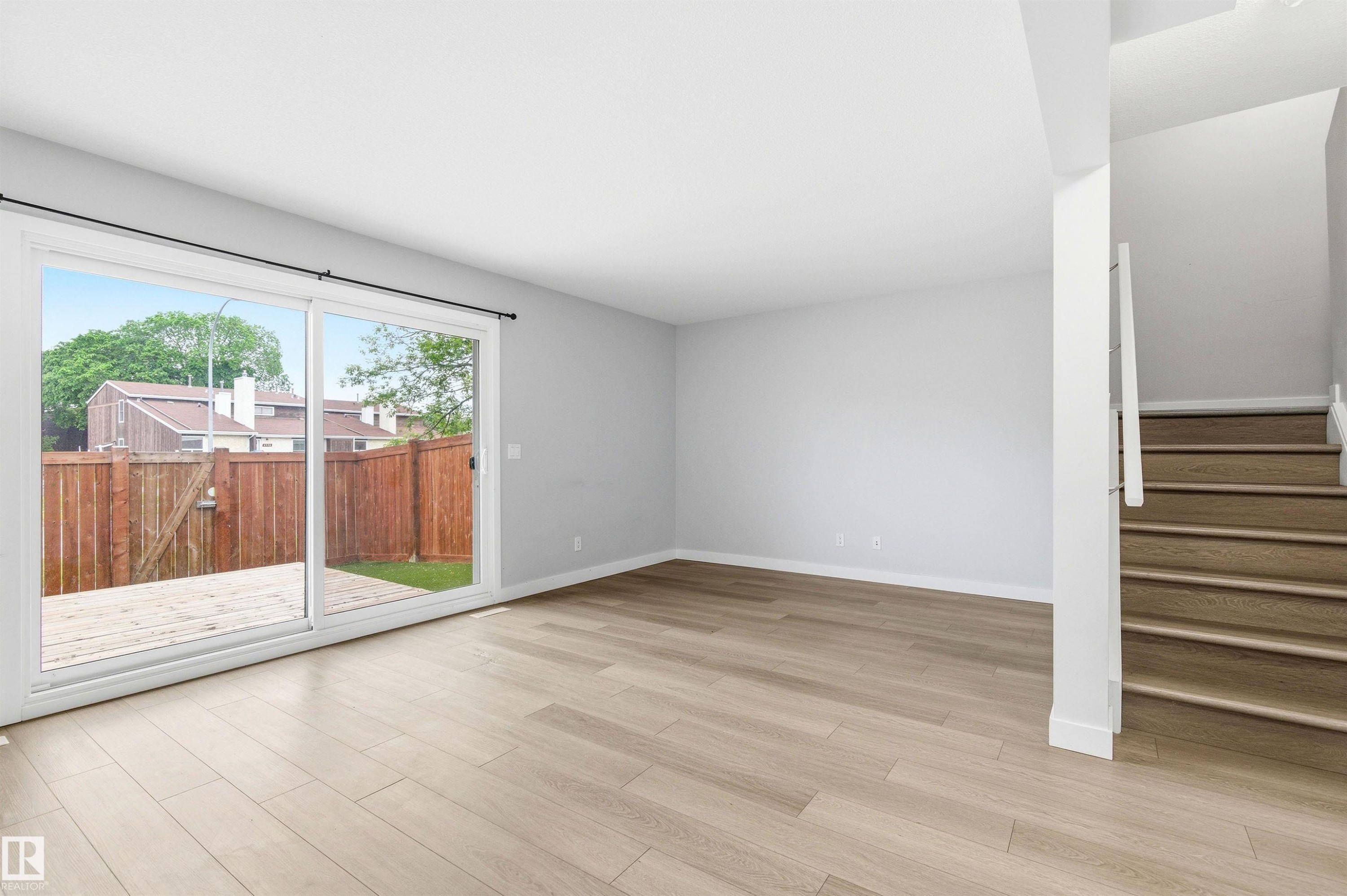 Spacious main living area featuring light wood-finish flooring, neutral paint, a large glass sliding door, and a staircase with contemporary railing - 18353 66 Avenue, Edmonton, AB