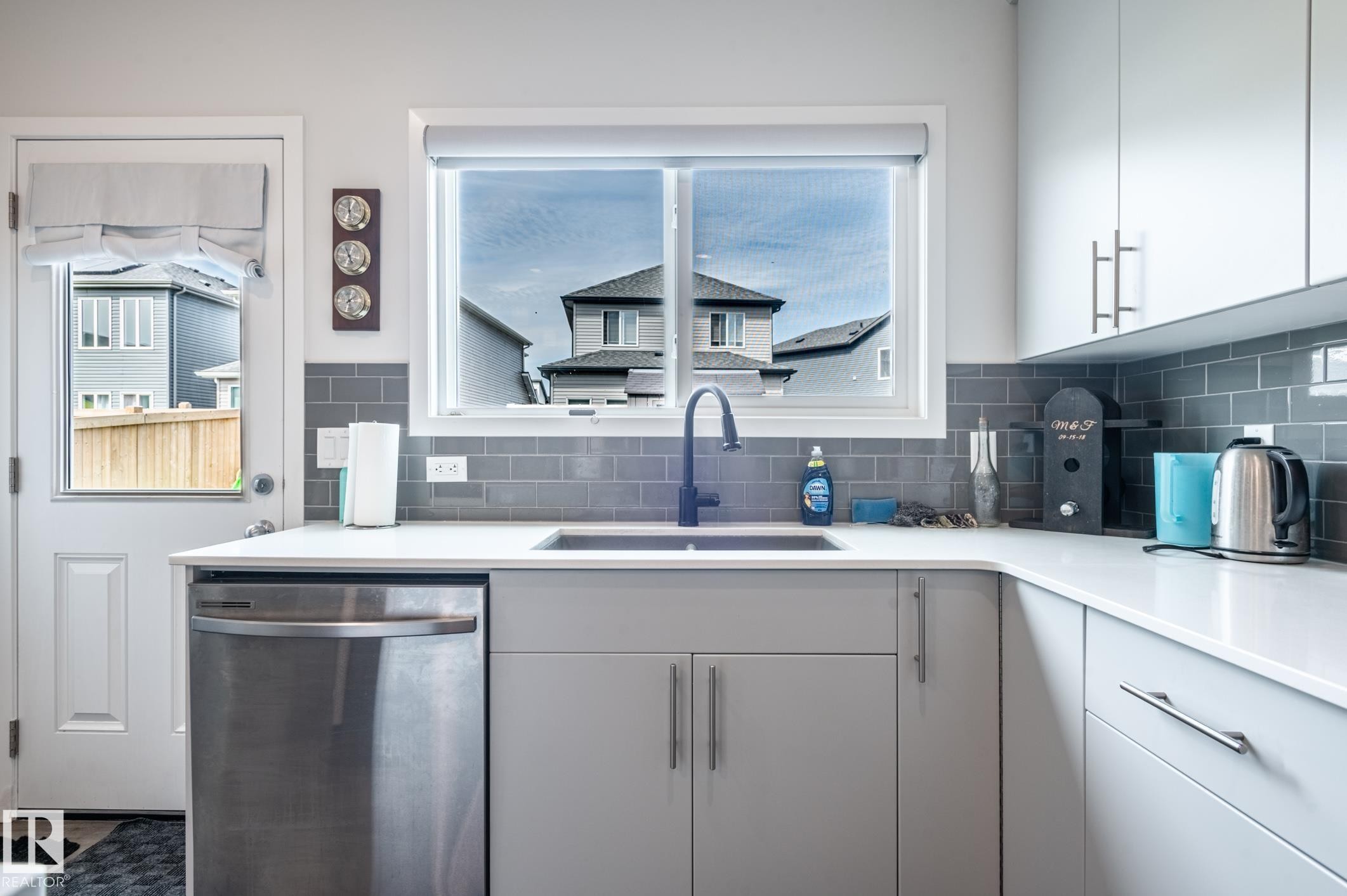 Modern kitchen featuring light gray cabinetry with brushed nickel hardware, white countertops, a stainless steel dishwasher, a single basin sink with a matte black faucet, and a gray subway tile backsplash - 17562 63A Street, Edmonton, AB