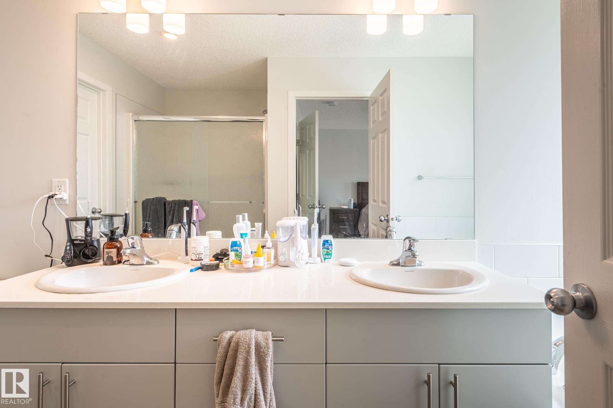Dual undermount sinks set into a light-toned countertop, complemented by a large vanity mirror and upper light fixture - 17562 63A Street, Edmonton, AB