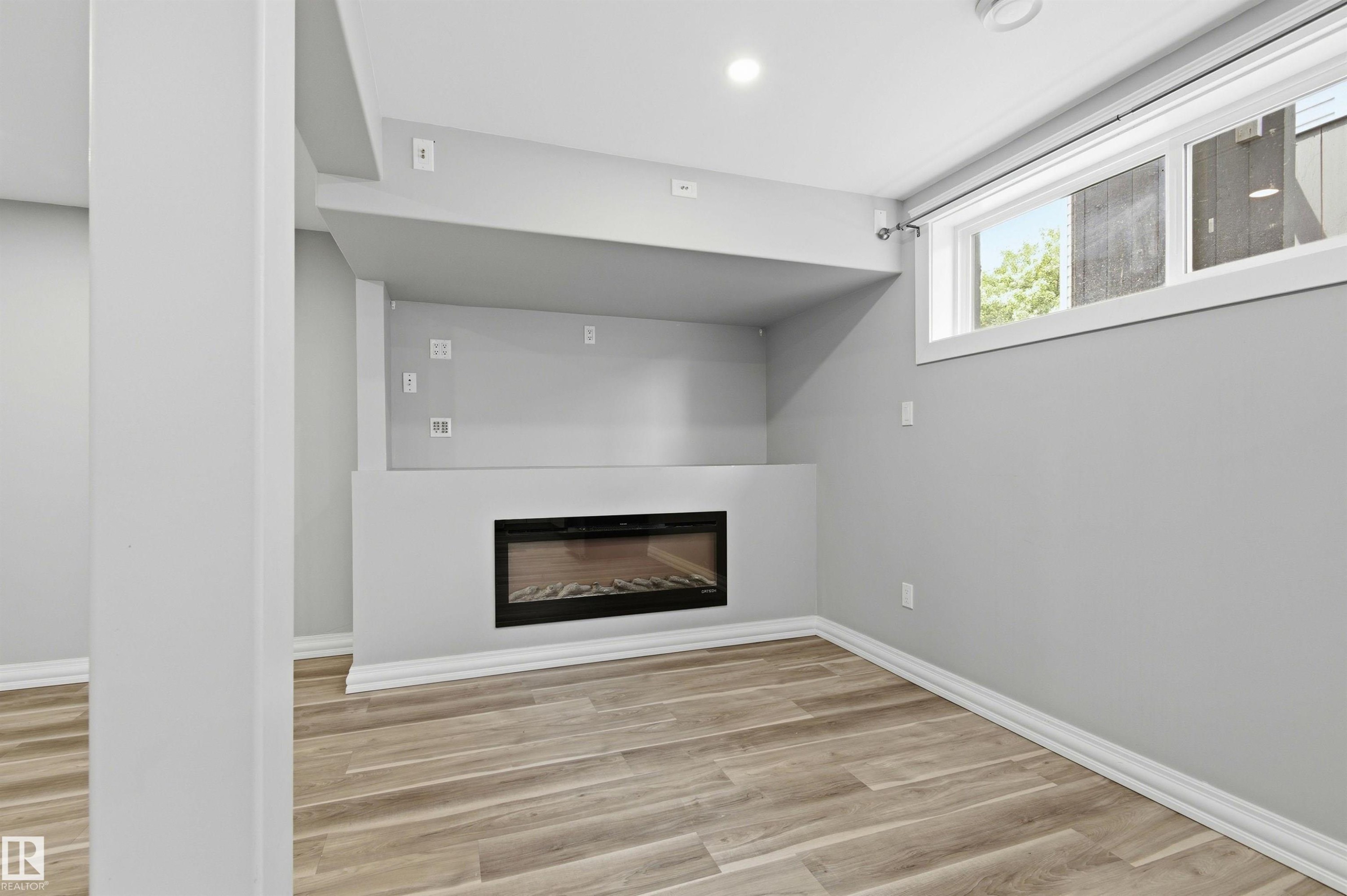 Recessed electric fireplace with a built-in media niche, wood-finish flooring, and a large egress window - 7412/7414 83 Avenue Nw, Edmonton, AB - Indoor With Fireplace