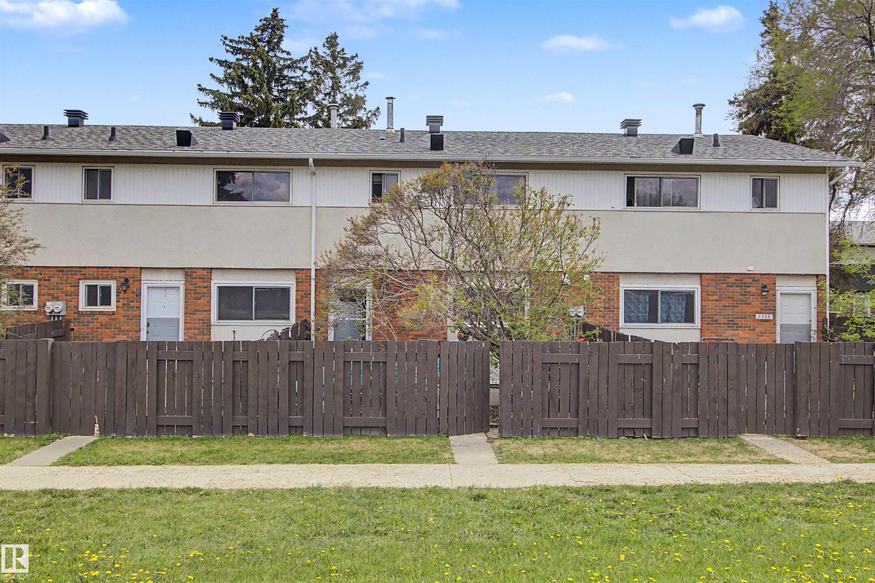 Multi-story townhome with a brick and stucco facade, featuring a wood fence, mature landscaping, and a sidewalk - 5724 143 Avenue, Edmonton, AB