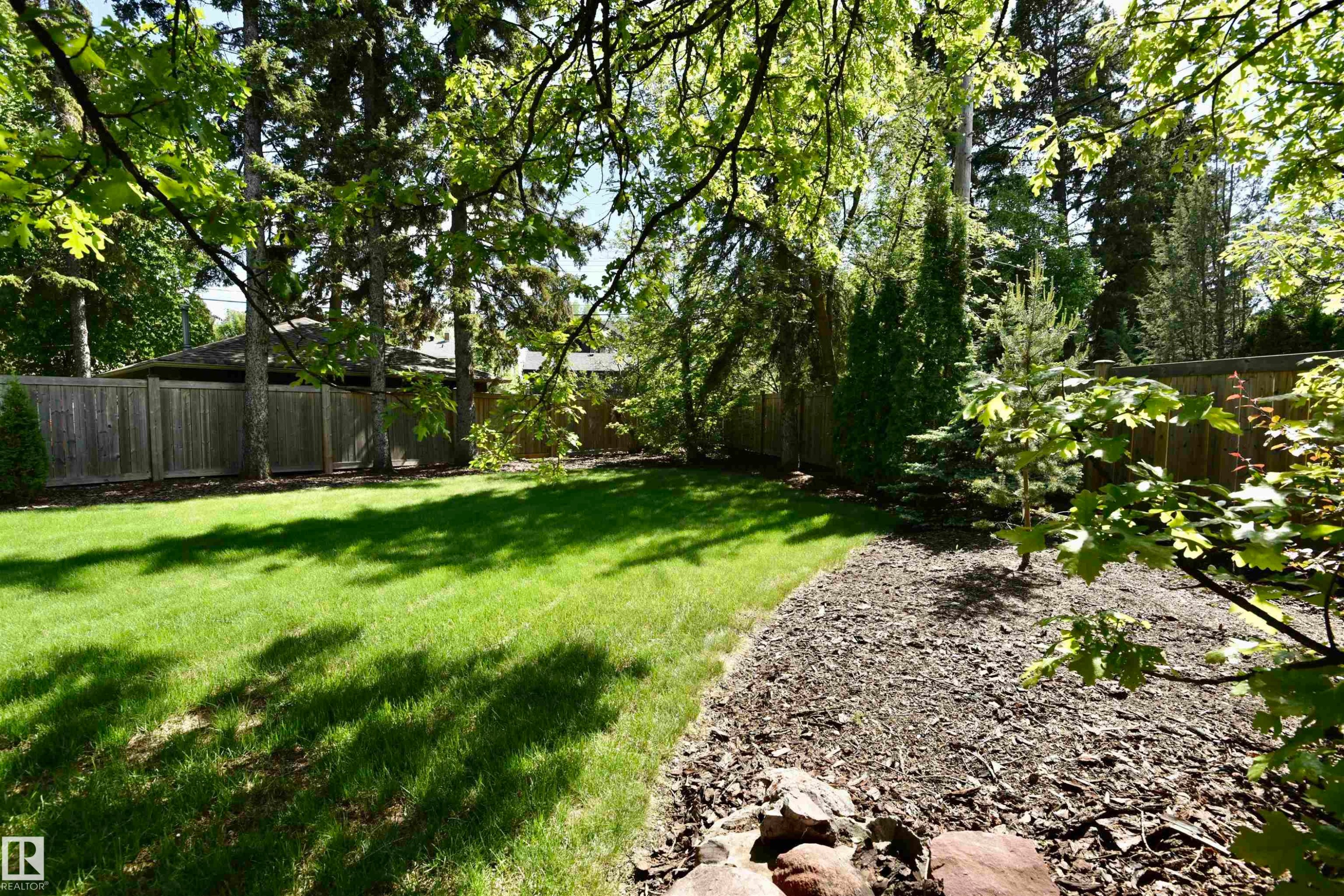 Expansive backyard featuring a manicured lawn, established perimeter fencing, mature trees, a landscaped bark-mulch bed, and natural rock accents - 6 St George'S Crescent, Edmonton, AB