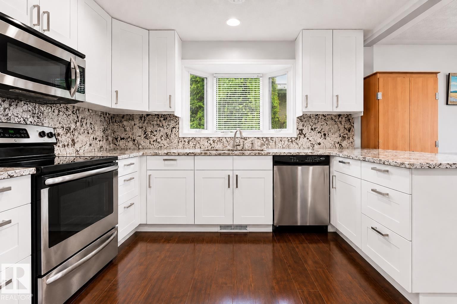 U-shaped kitchen featuring white cabinetry, stainless steel appliances, speckled countertops, a bay window, and wood-finish flooring - 11234 40 Avenue Nw, Edmonton, AB - Indoor Photo Showing Kitchen With Upgraded Kitchen