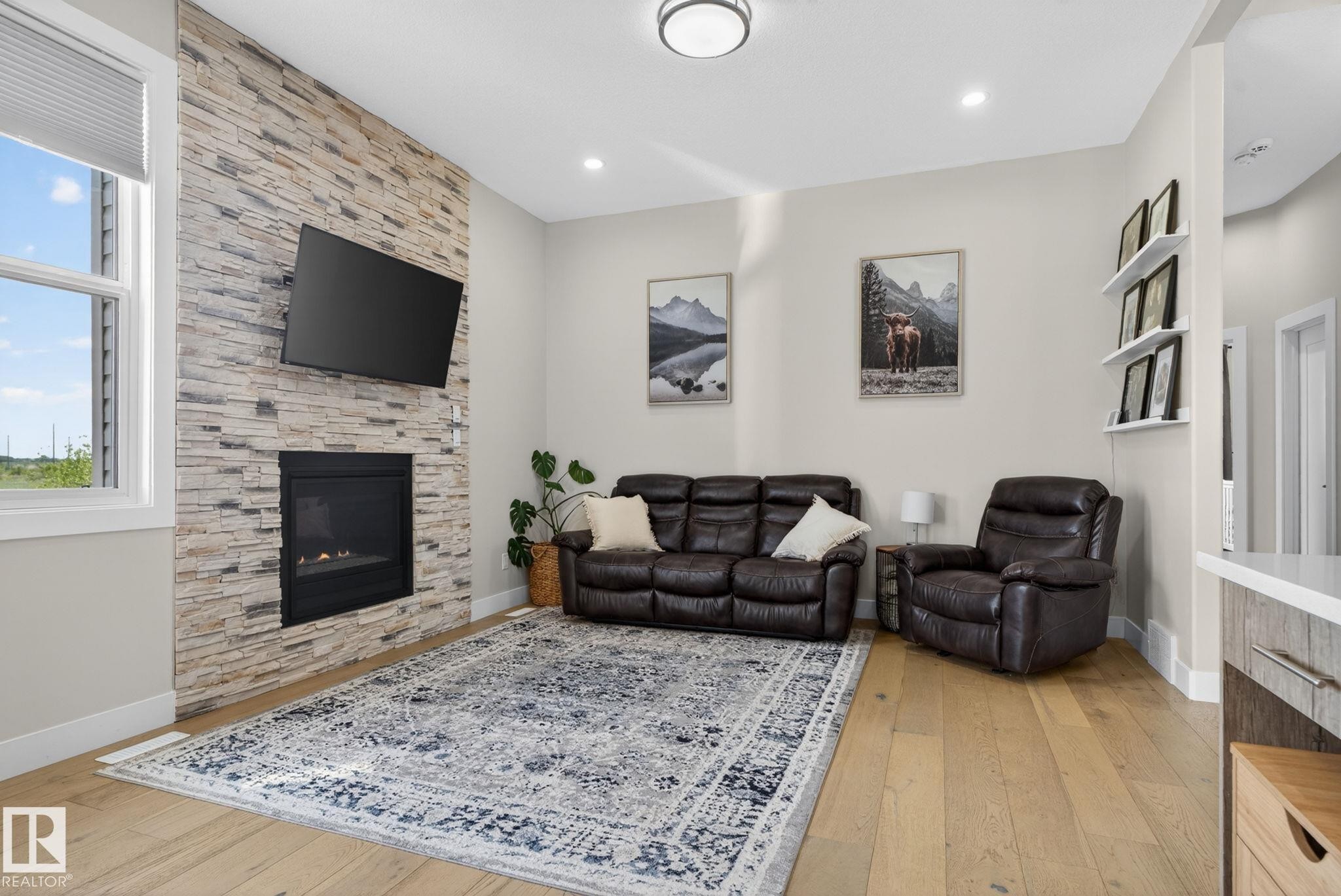 Living area featuring a floor-to-ceiling stacked stone fireplace, a large picture window, recessed lighting, wood-finish flooring, and built-in floating shelves - 4204 55Av, Lamont, AB