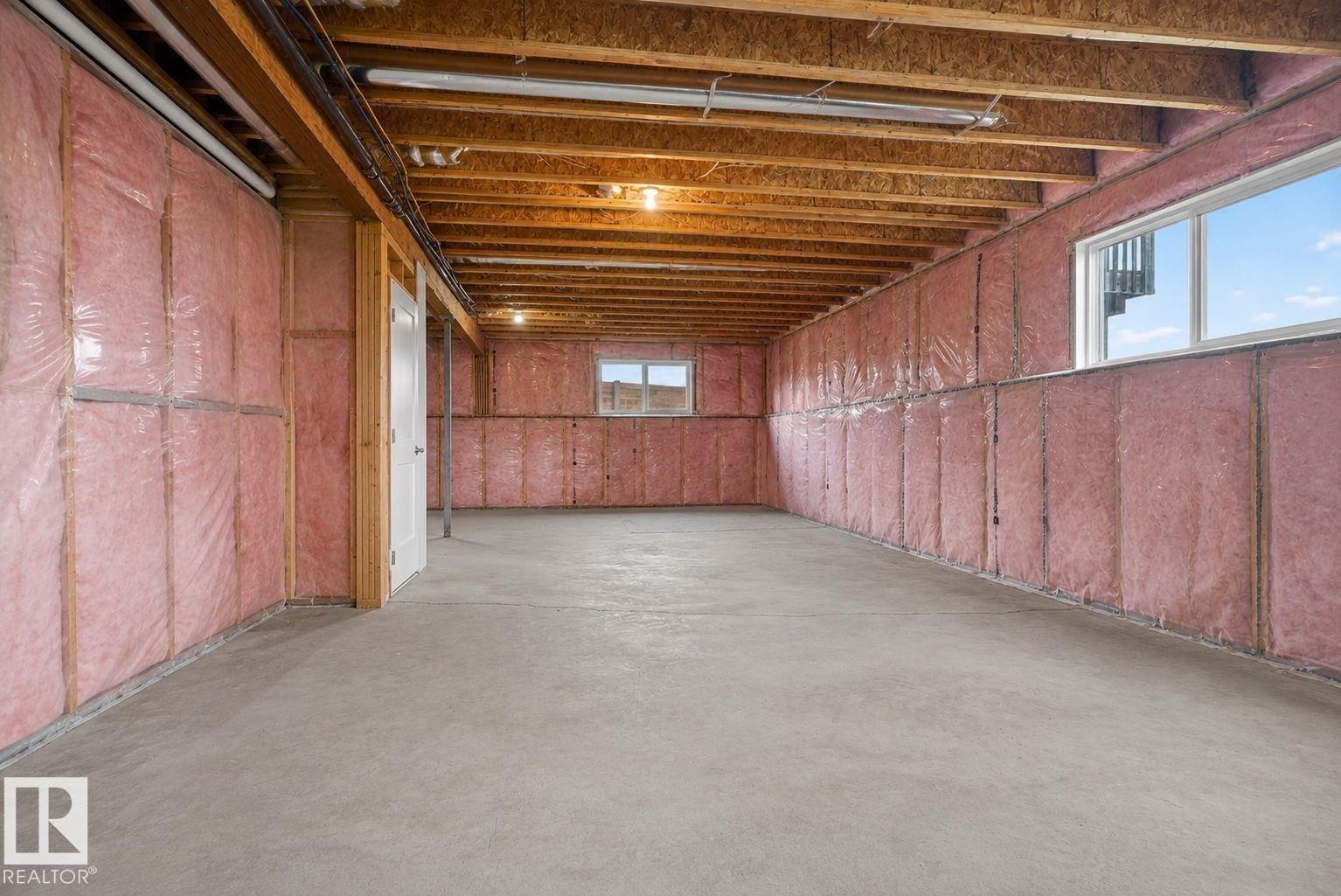 Unfinished lower level with concrete flooring, exposed wood joists, framed walls with pink insulation, two windows, and a white interior door - 4204 55Av, Lamont, AB