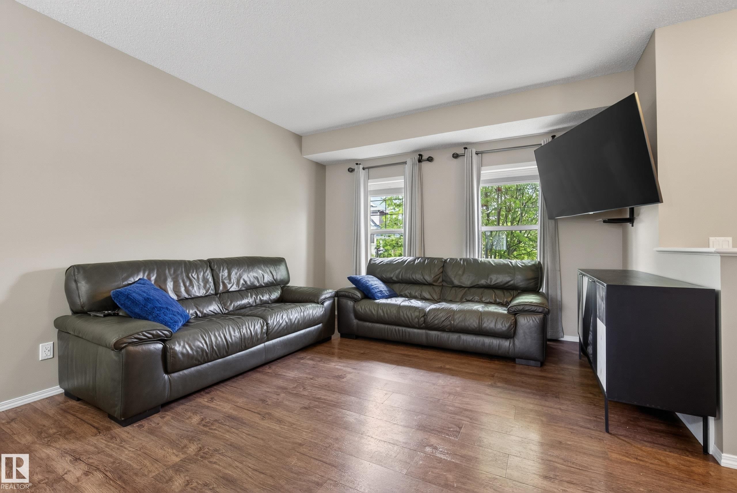 Living area featuring wood-finish flooring, neutral wall tones, and multiple windows with curtain rods - 2325 69A Street, Edmonton, AB