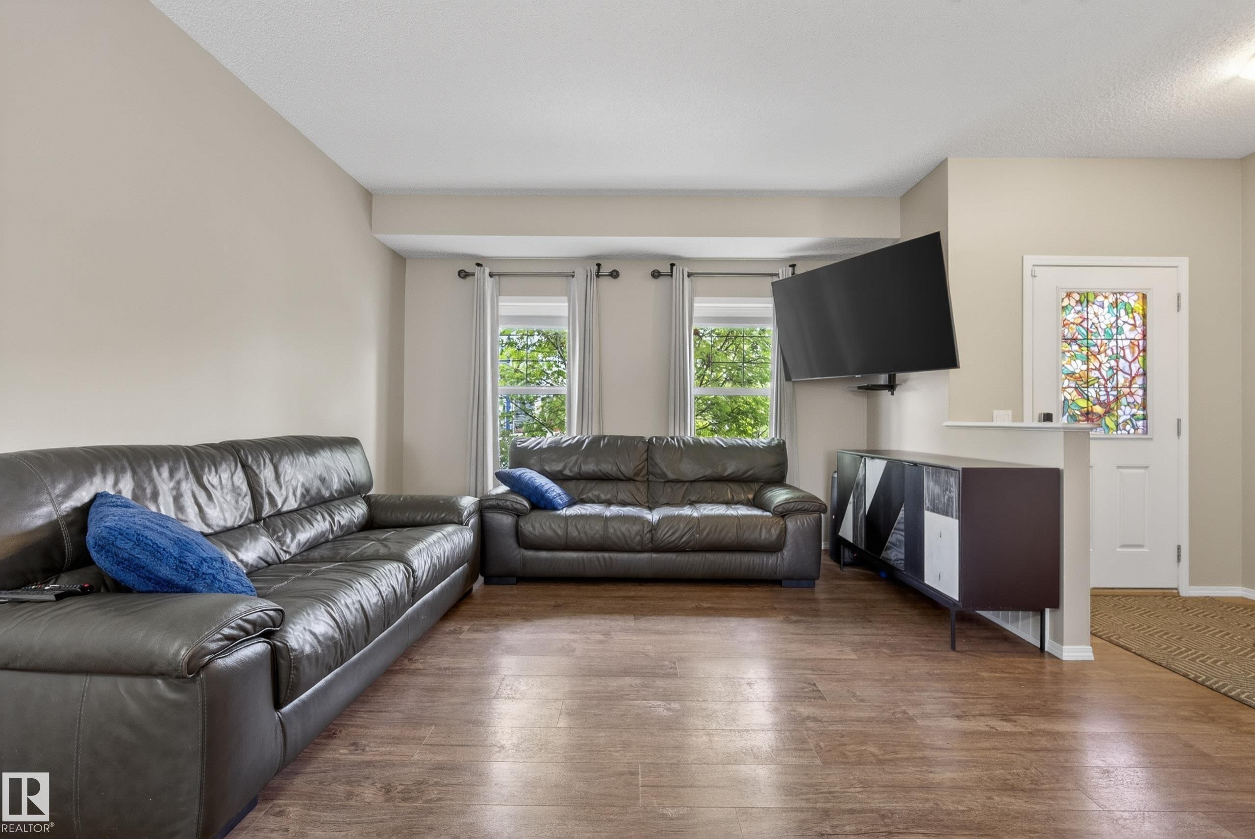 Spacious living area featuring wood-finish flooring, light-toned walls, a bay window with natural light, and a decorative front door with patterned glass - 2325 69A Street, Edmonton, AB