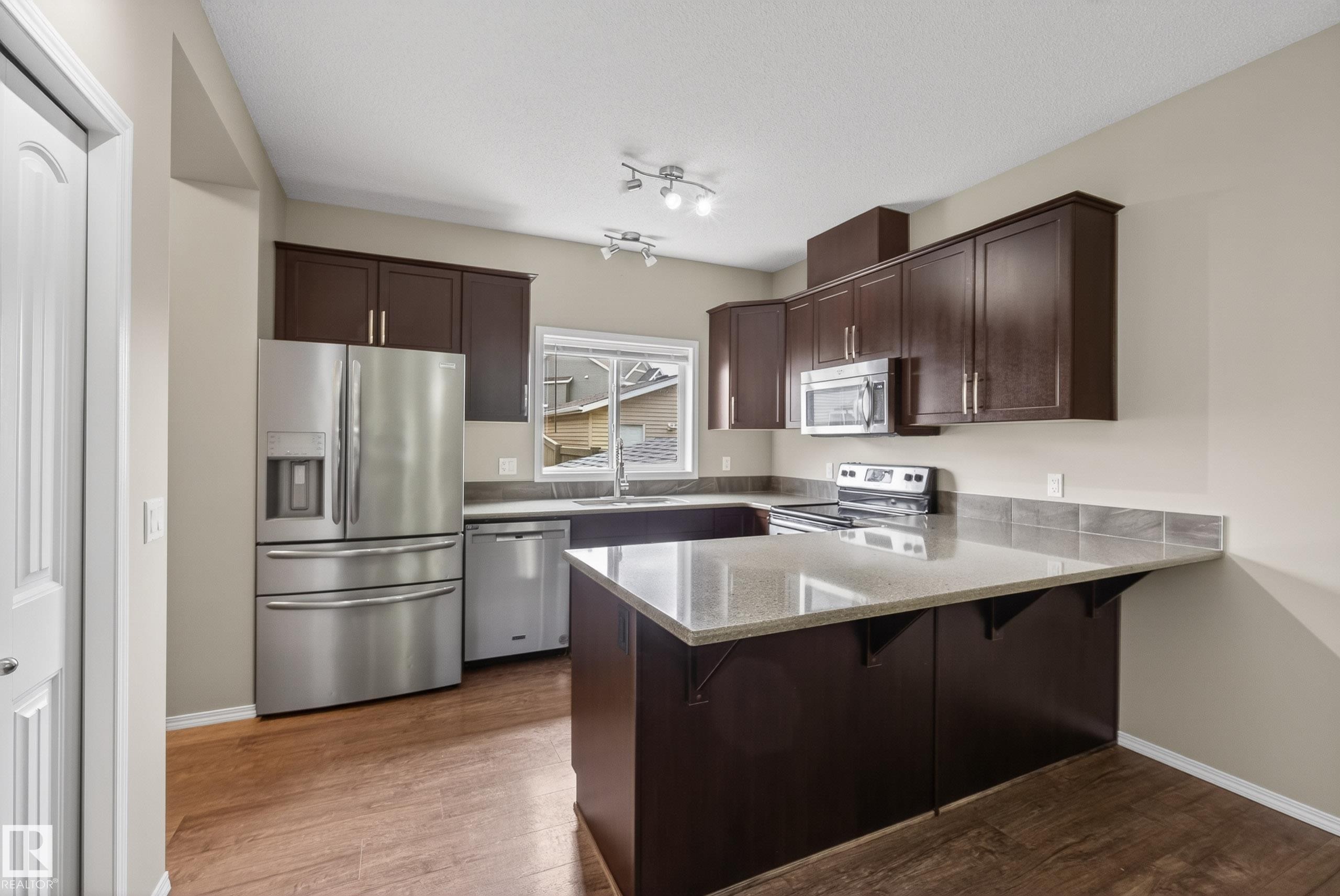 Kitchen featuring dark wood-finish cabinetry, stainless steel appliances, light-toned countertops, and wood-finish flooring - 2325 69A Street, Edmonton, AB