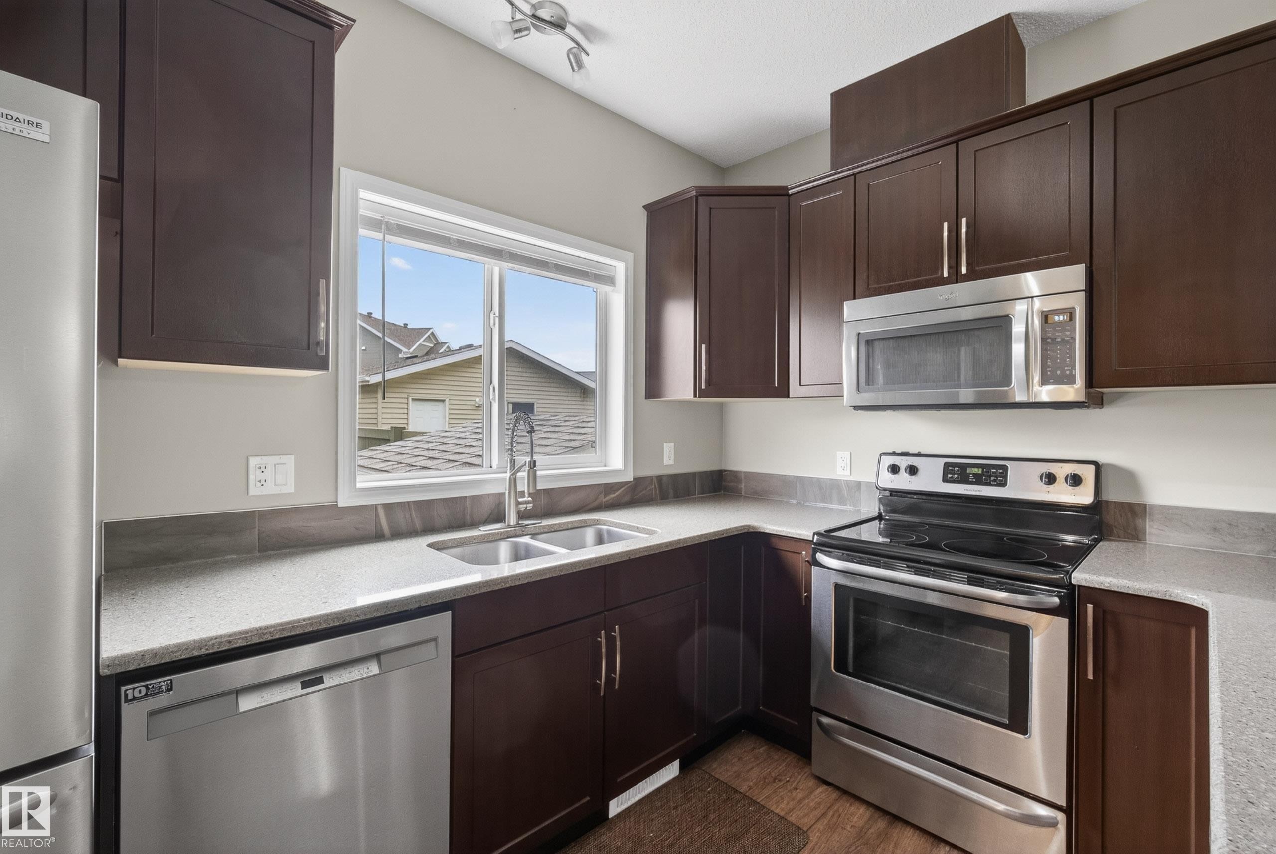 Kitchen featuring dark wood cabinetry, light-toned countertops, stainless steel appliances, and a double-basin sink with a window - 2325 69A Street, Edmonton, AB
