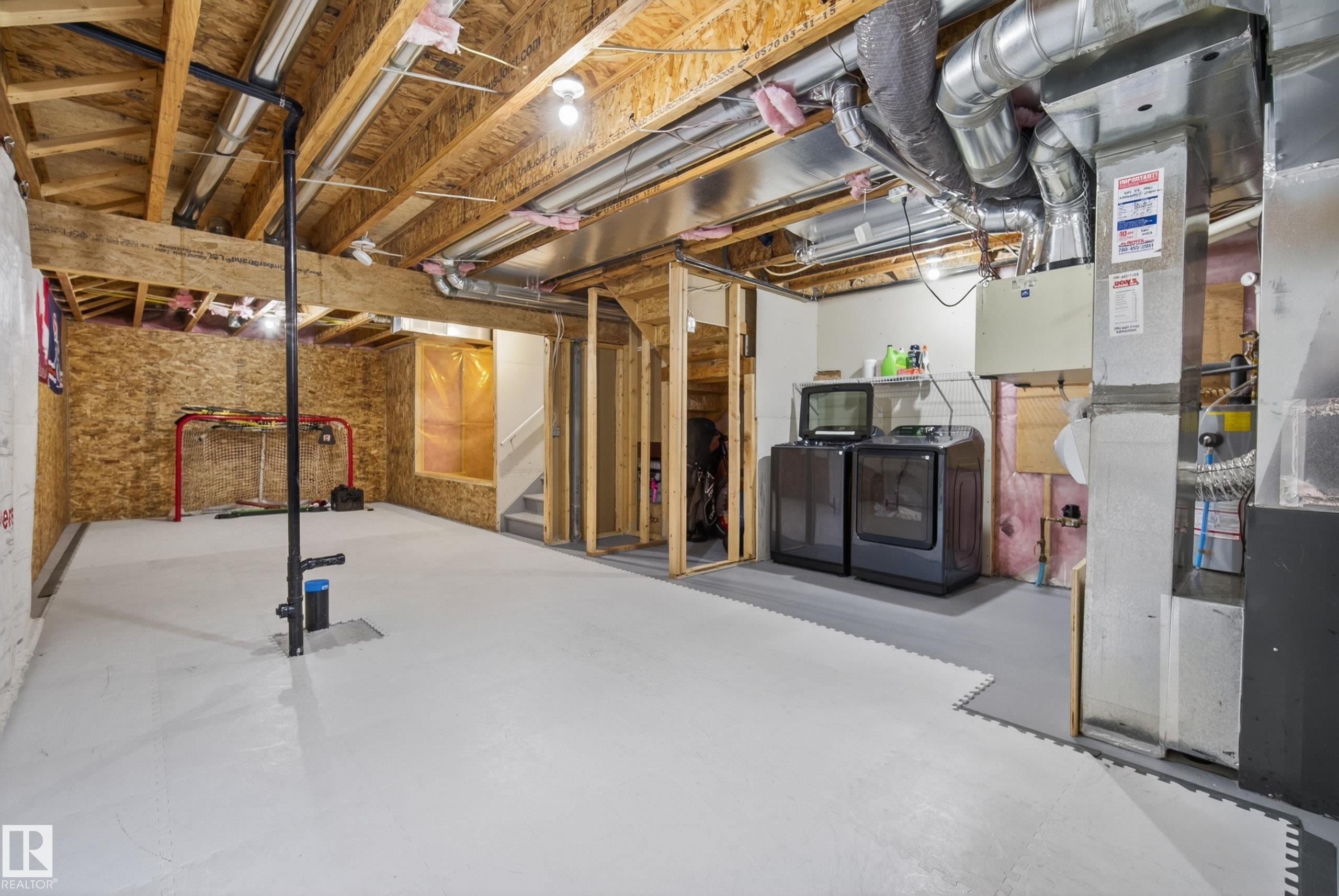 Spacious unfinished basement offering exposed wood joists and structural beams, painted concrete flooring, an accessible utility area with a washer and dryer, and a partially framed staircase - 2325 69A Street, Edmonton, AB