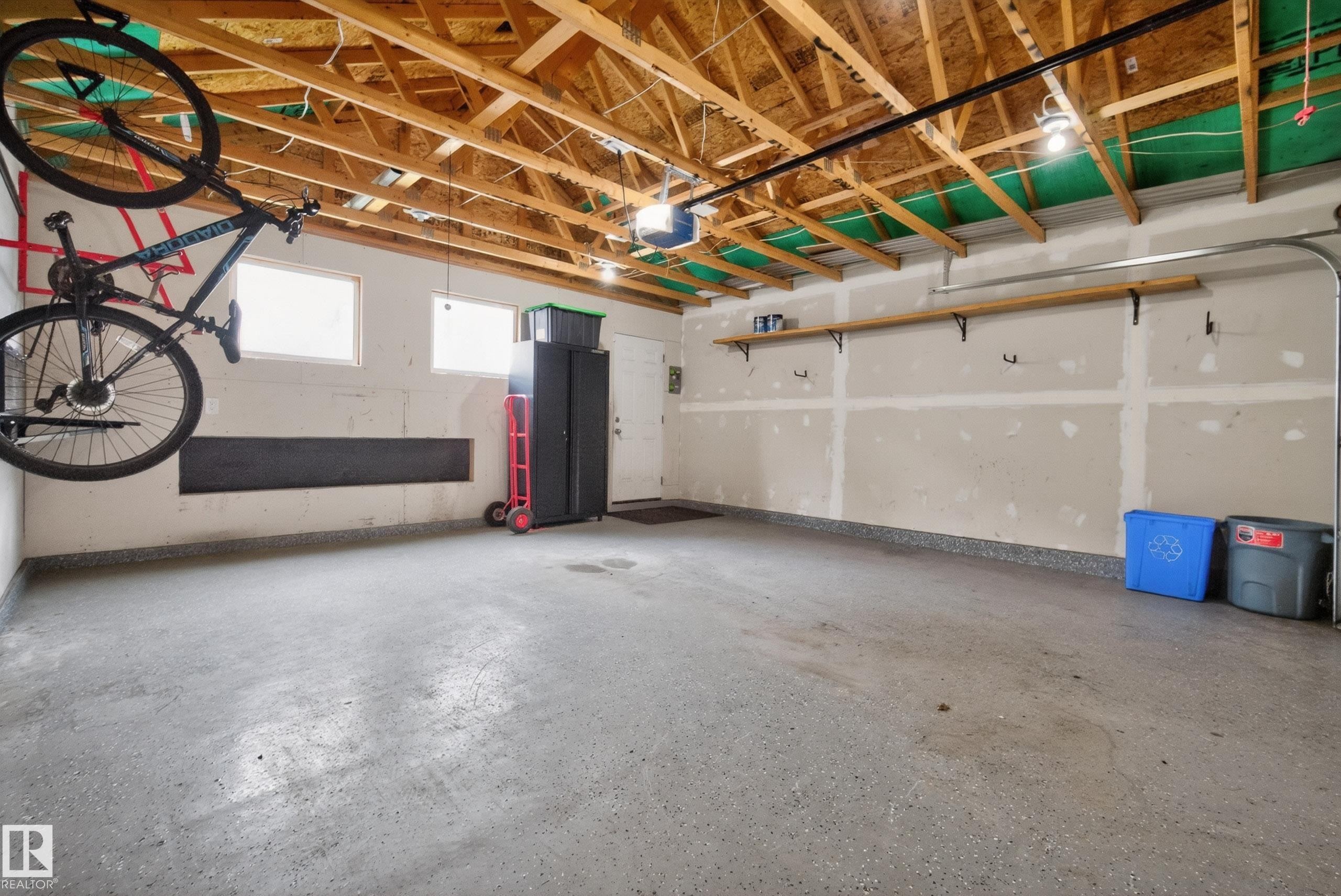 Spacious garage featuring an exposed beam ceiling, concrete flooring, upper wall shelving, and multiple windows providing natural light - 2325 69A Street, Edmonton, AB