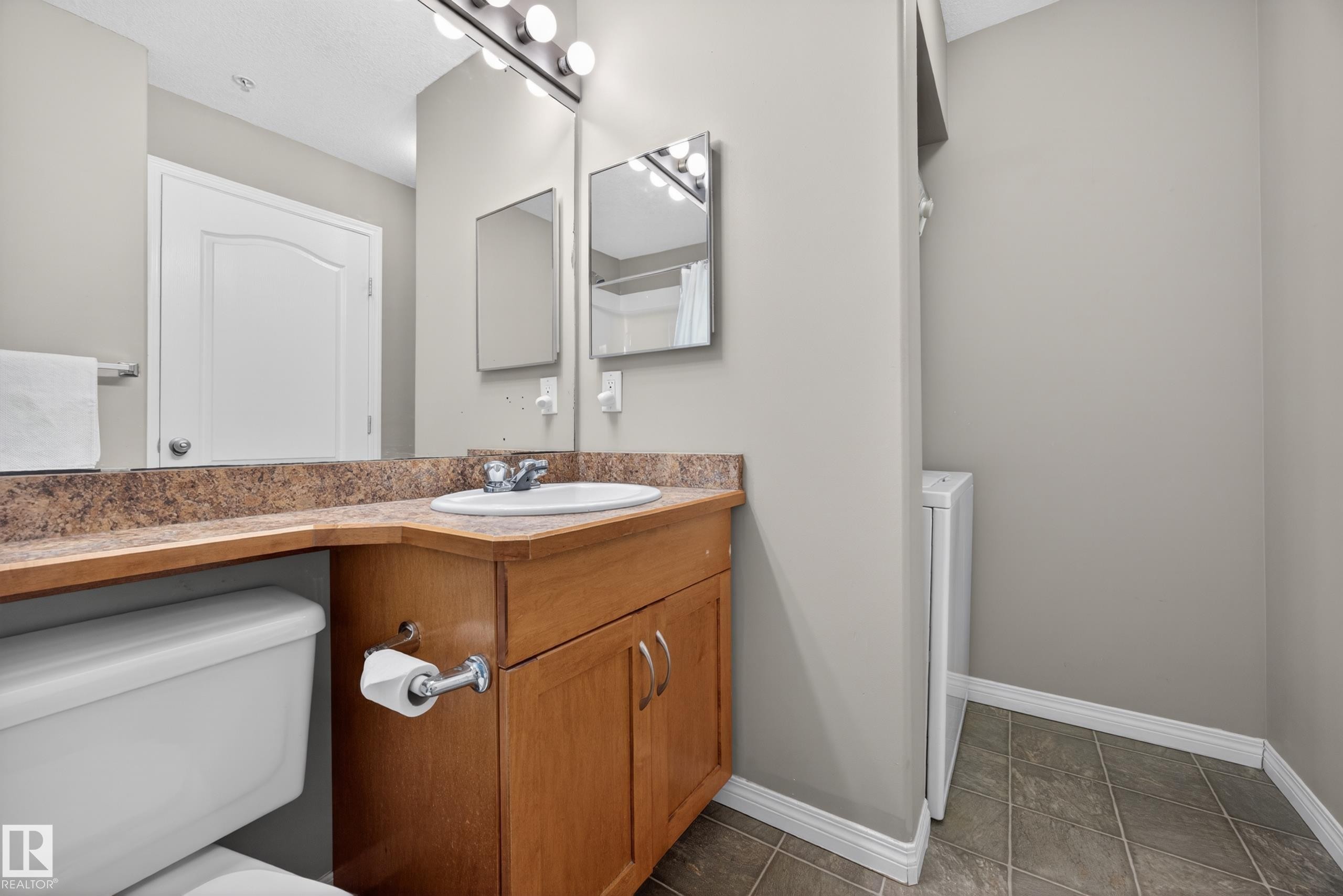 Bathroom featuring a light wood-finish vanity with a brown speckled countertop, an integrated sink, and a mirrored wall - 111 3425 19, Edmonton, AB