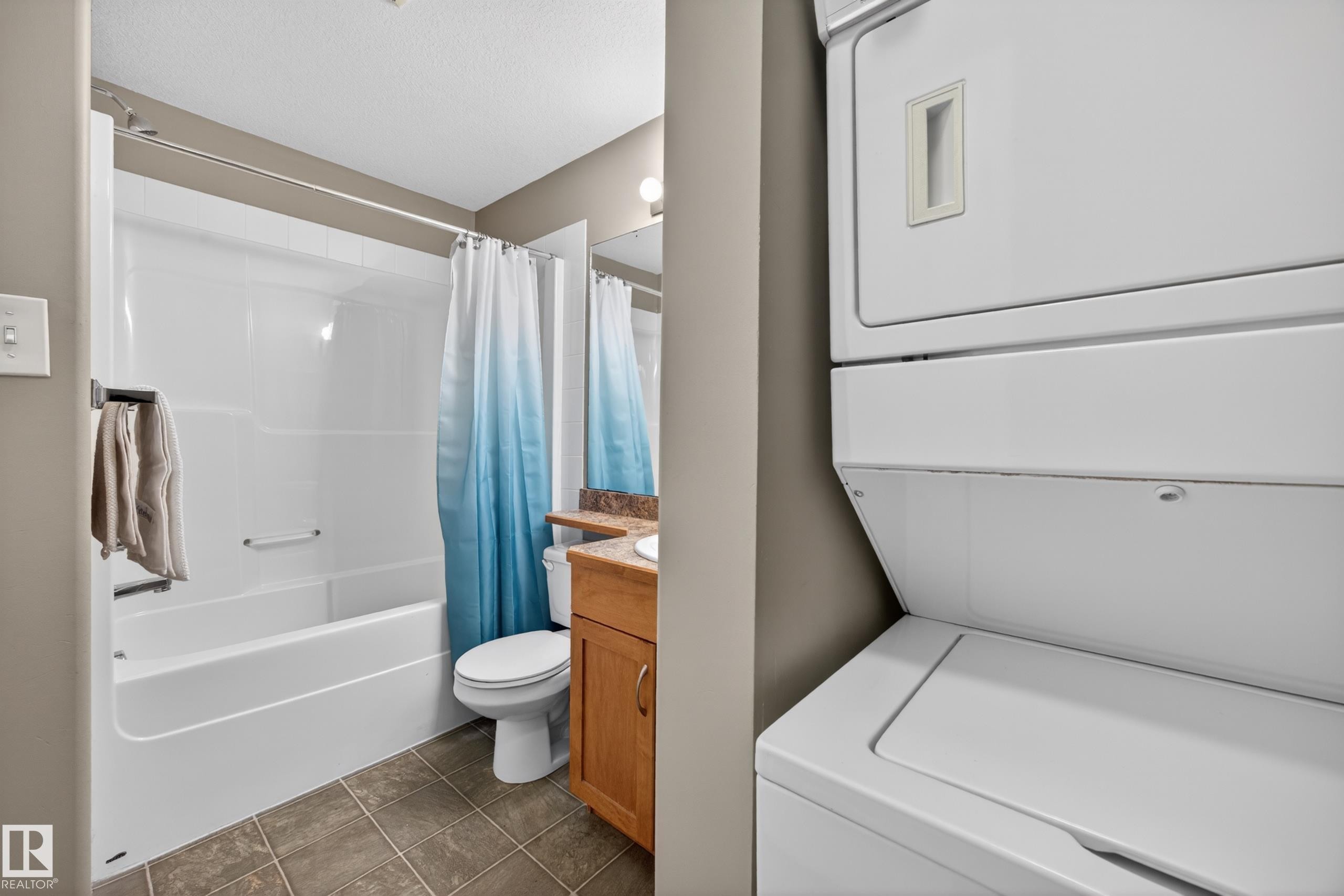 Full bathroom featuring a white tub and shower combination with a single basin vanity, brown wood cabinetry, and a stacked washer and dryer - 111 3425 19, Edmonton, AB