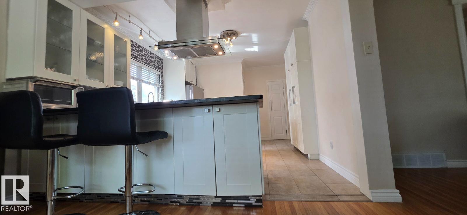 Kitchen featuring a central island with a dark countertop and integrated cabinetry, a stainless steel range hood, glass-front upper cabinets, and a tile backsplash - 6704 93 Street, Edmonton, AB
