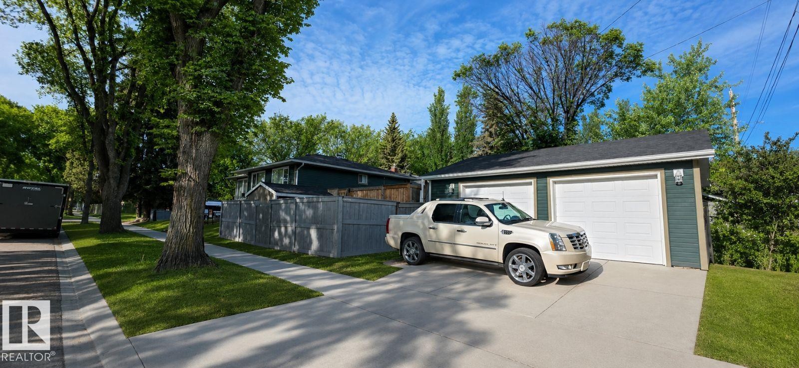 Detached garage with dual white garage doors and a paved driveway, set against a backdrop of mature trees and established landscaping - 6704 93 Street, Edmonton, AB