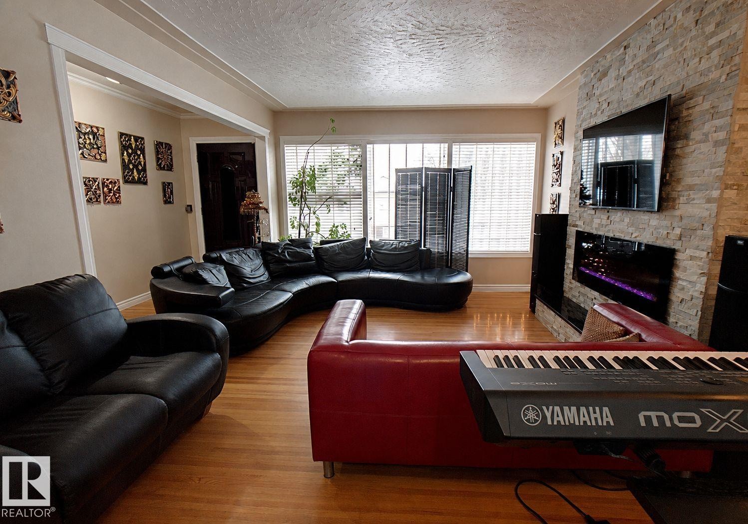 Living area featuring wood-finish flooring, a stone-accented fireplace, and a large window with horizontal blinds - 6704 93 Street, Edmonton, AB