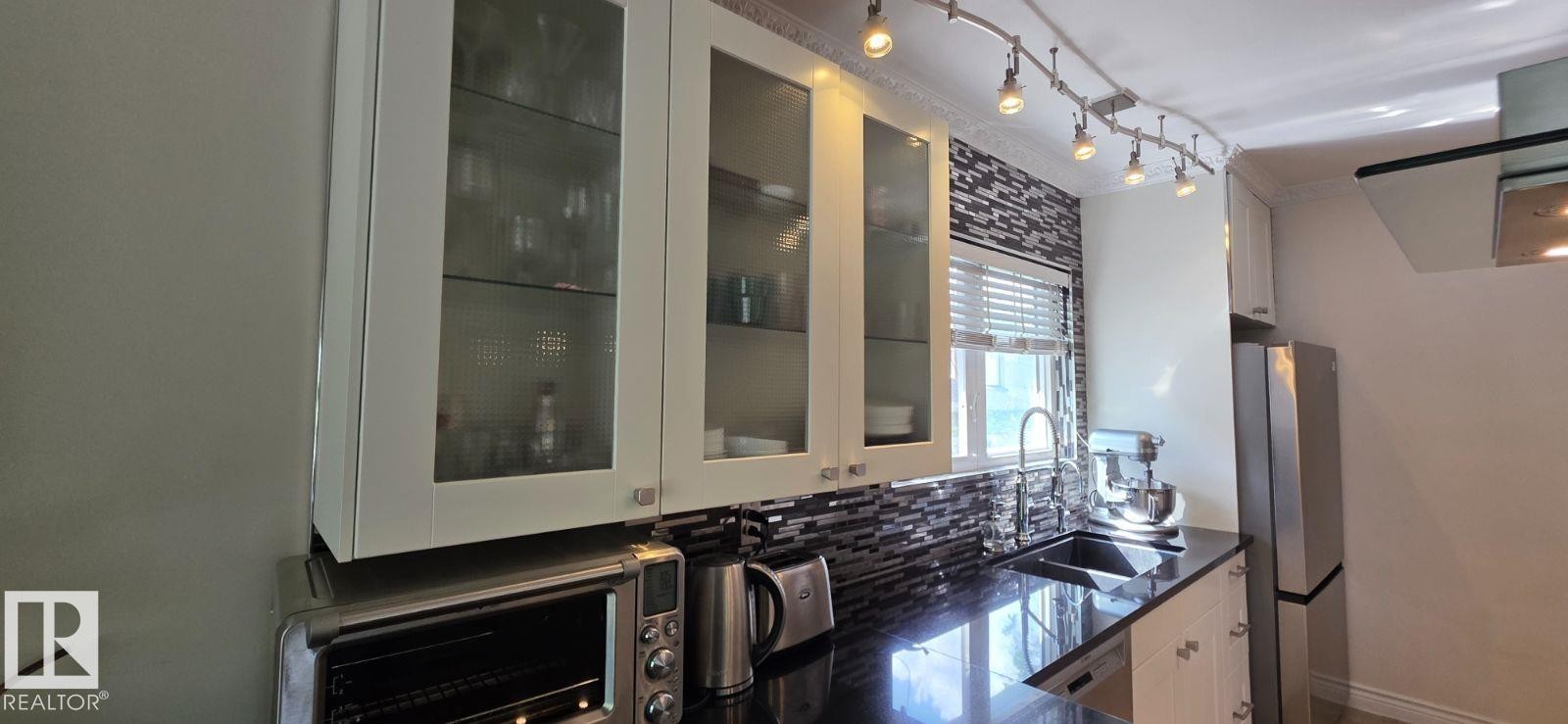 Kitchen area featuring white cabinetry with glass inserts, a black granite-style countertop, and a mosaic tile backsplash - 6704 93 Street, Edmonton, AB