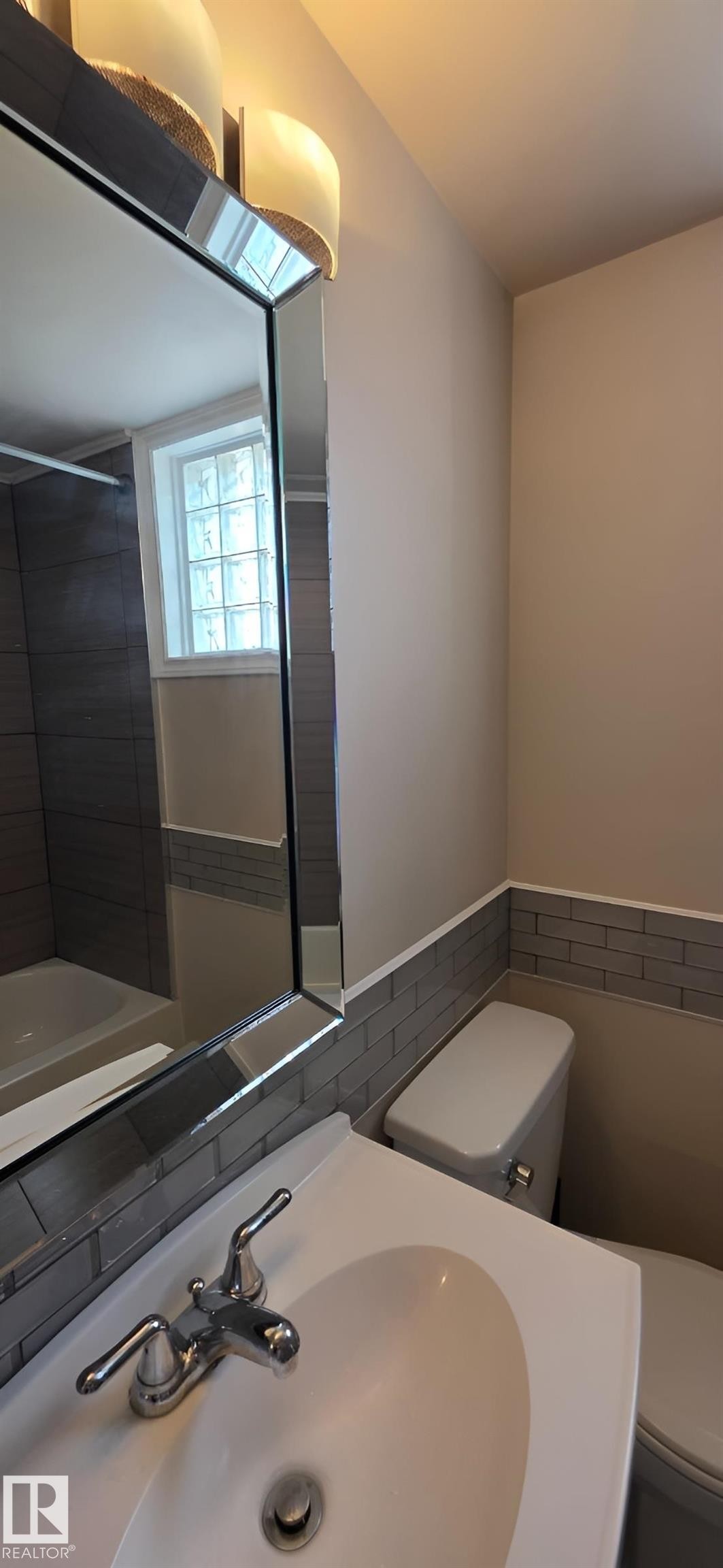 Bathroom featuring a porcelain sink with chrome fixtures, a toilet, a decorative mirror, and a window with glass block accents - 6704 93 Street, Edmonton, AB