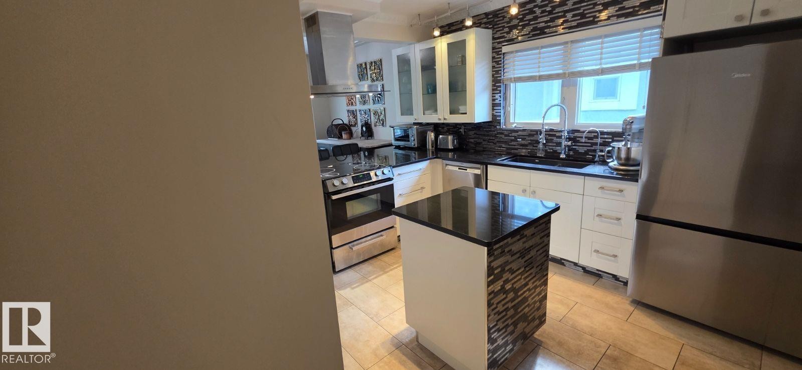 Contemporary kitchen featuring white cabinetry, dark countertops, a tiled backsplash, stainless steel appliances, and a central island with a contrasting stone-look finish - 6704 93 Street, Edmonton, AB