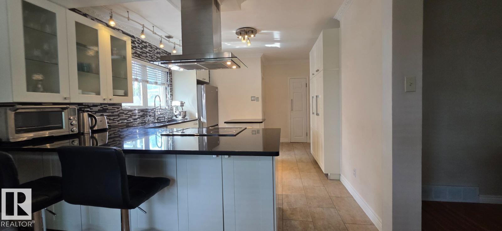 Contemporary kitchen featuring an island with built-in stovetop, black countertops, stainless steel range hood, glass-front upper cabinetry, and a tile backsplash - 6704 93 Street, Edmonton, AB