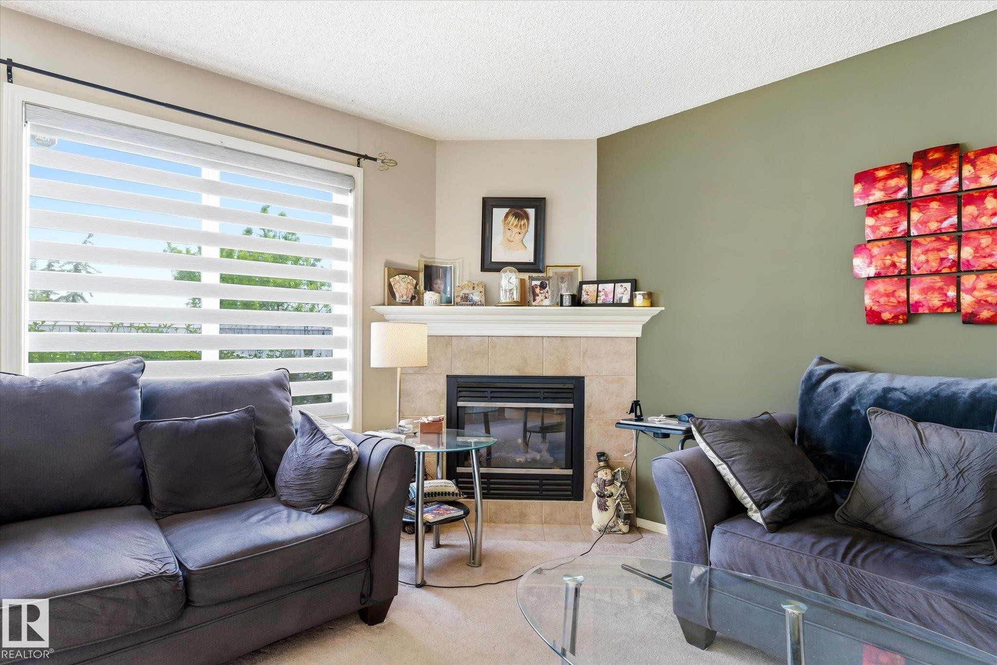 Living area featuring a tiled fireplace with mantel, large window with horizontal blinds, and neutral-toned wall paint - #37 4020 21 St Nw, Edmonton, AB - Indoor Photo Showing Living Room With Fireplace
